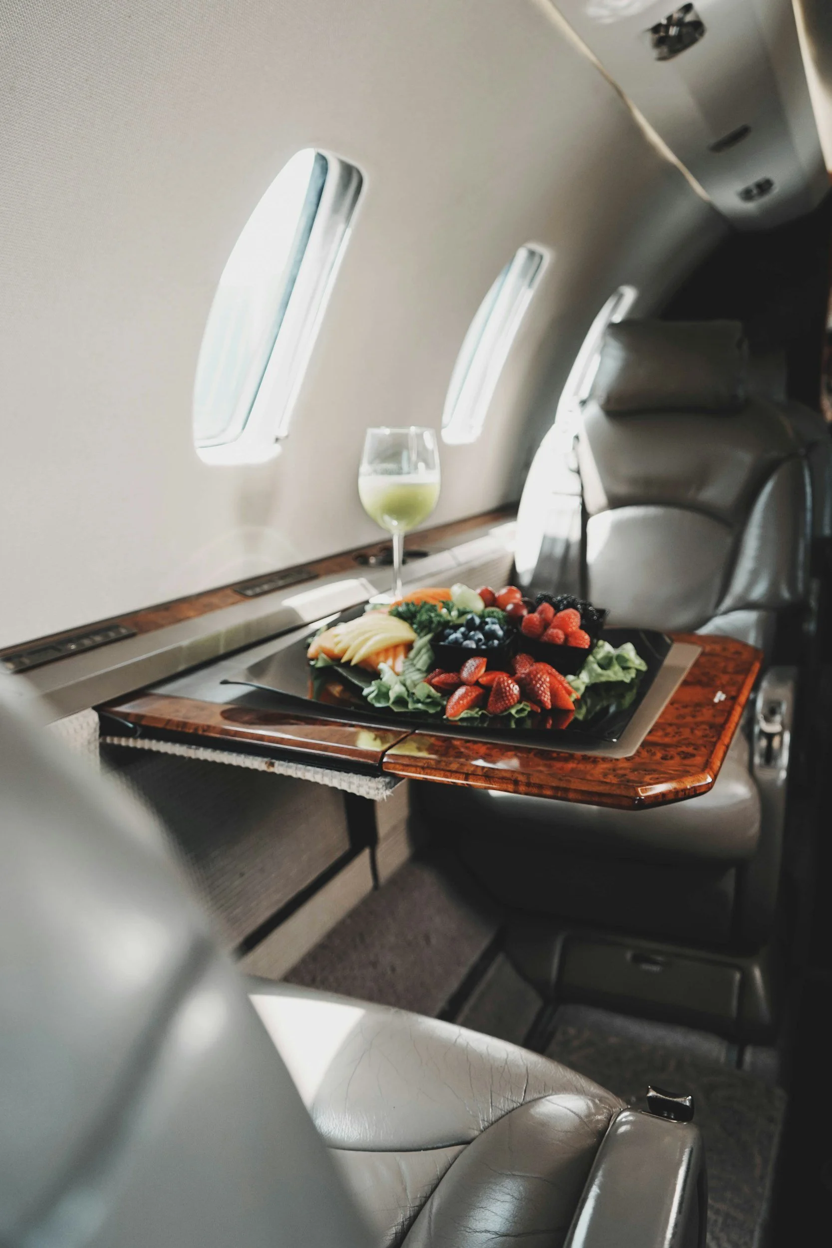 Photo of the interior of an airplane cabin showing a row of beige leather seats, a tray table with a tray of fresh fruit, and a glass of green drink. The ceiling has three rectangular overhead lights and air vents.