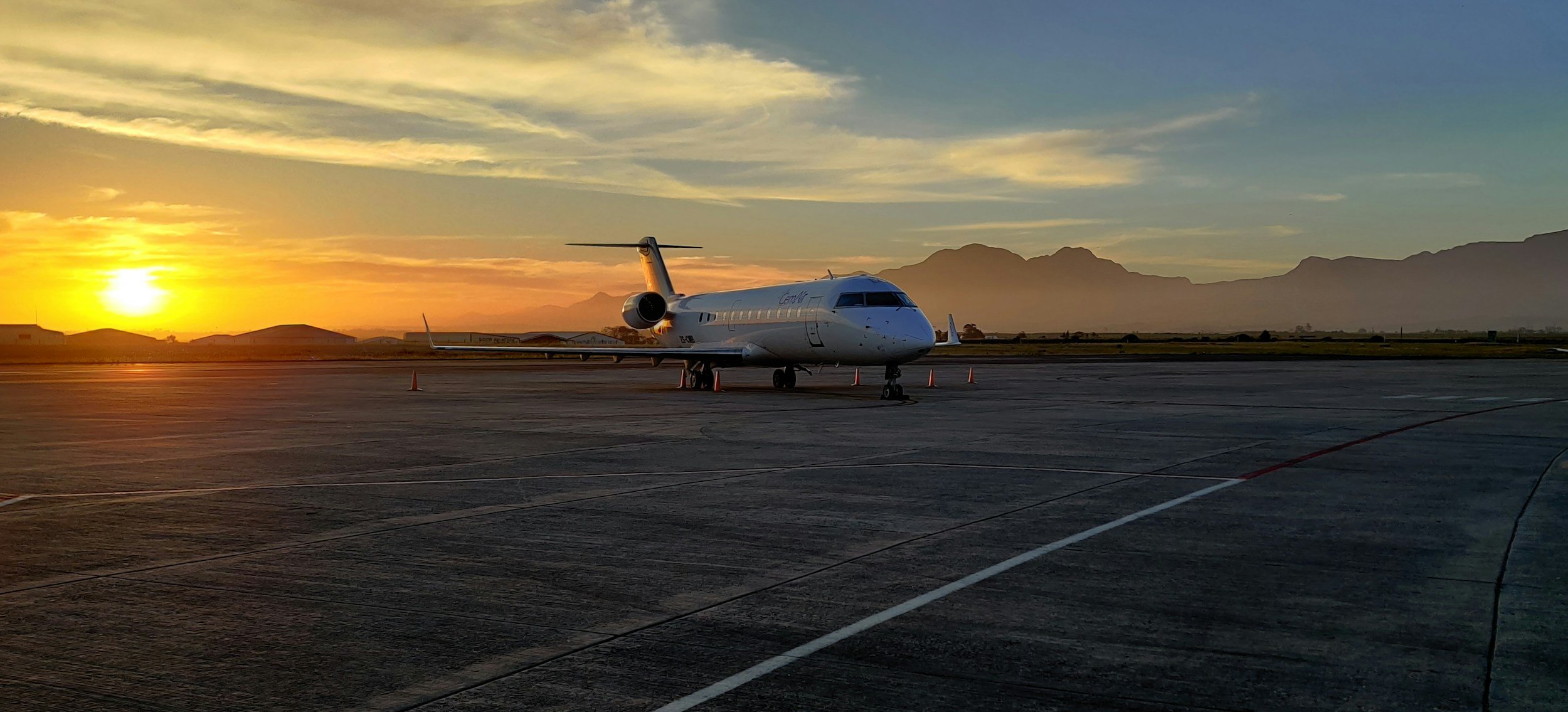A white private jet parked on an airport tarmac during sunset, with mountains in the background and a partly cloudy sky.