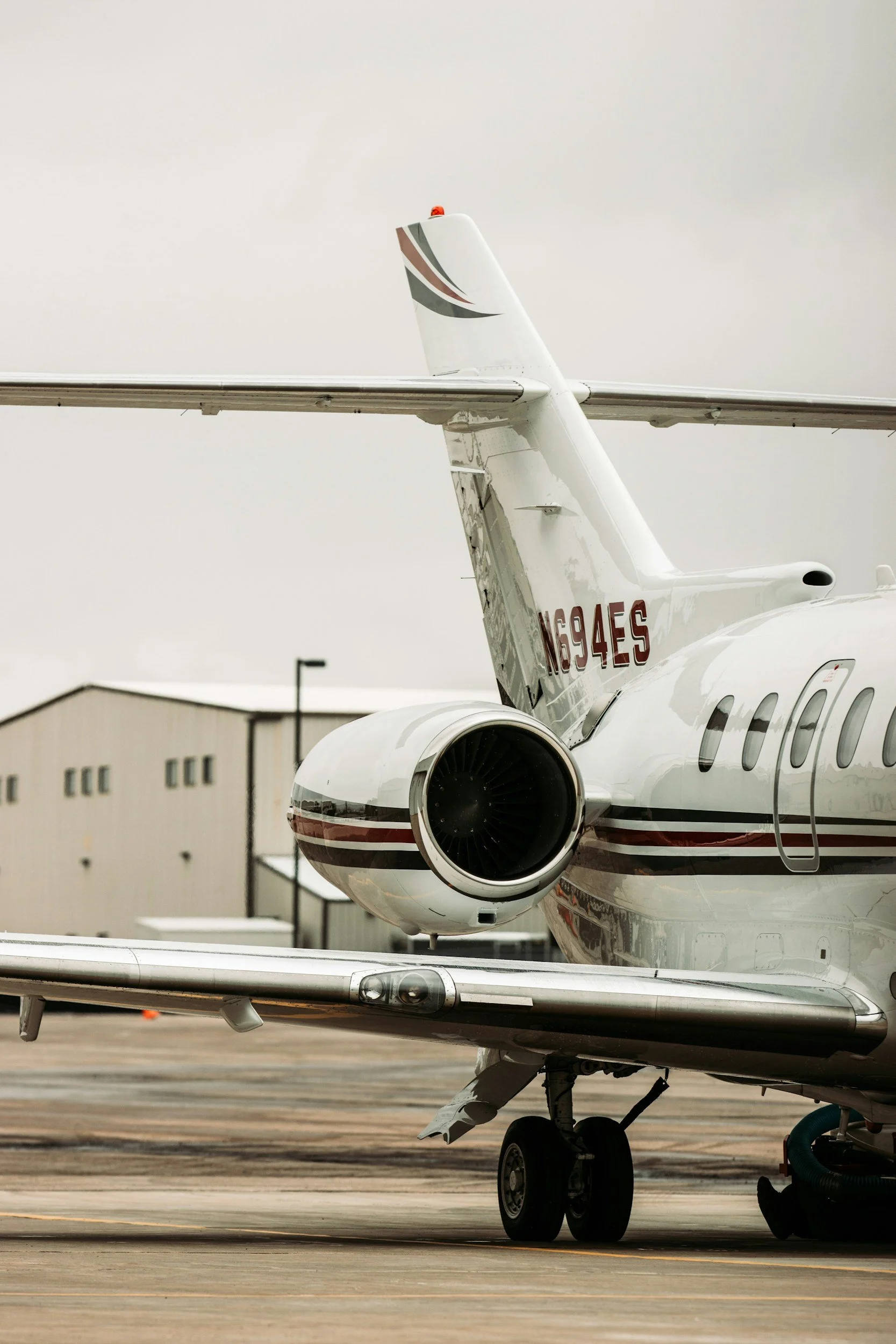 Close-up of a private jet on the tarmac, showing the tail with an airline logo, part of the fuselage, and engine with landing gear visible.