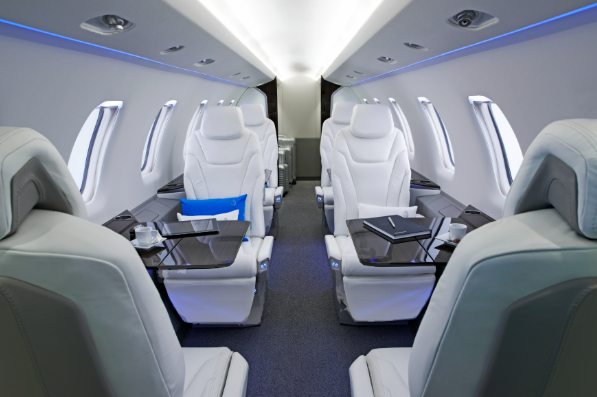Luxury airplane cabin with white leather seats and blue accent lighting, arranged in pairs with personal trays, glasses, and notebooks on the tables.
