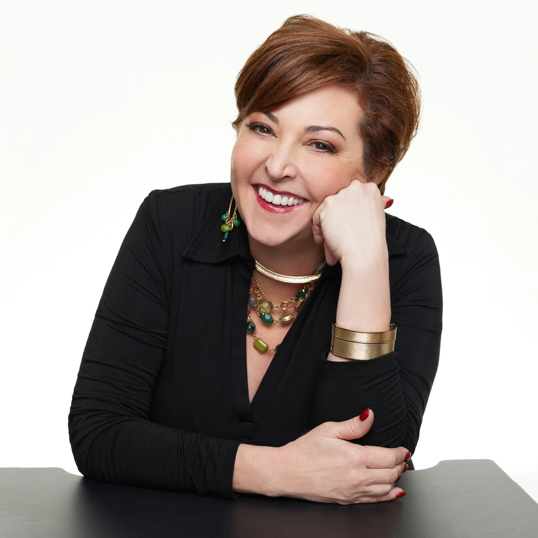 A smiling woman with short red hair, wearing a black top, jewelry, and red nail polish, resting her chin on her hand with folded arms on a dark surface against a plain white background.