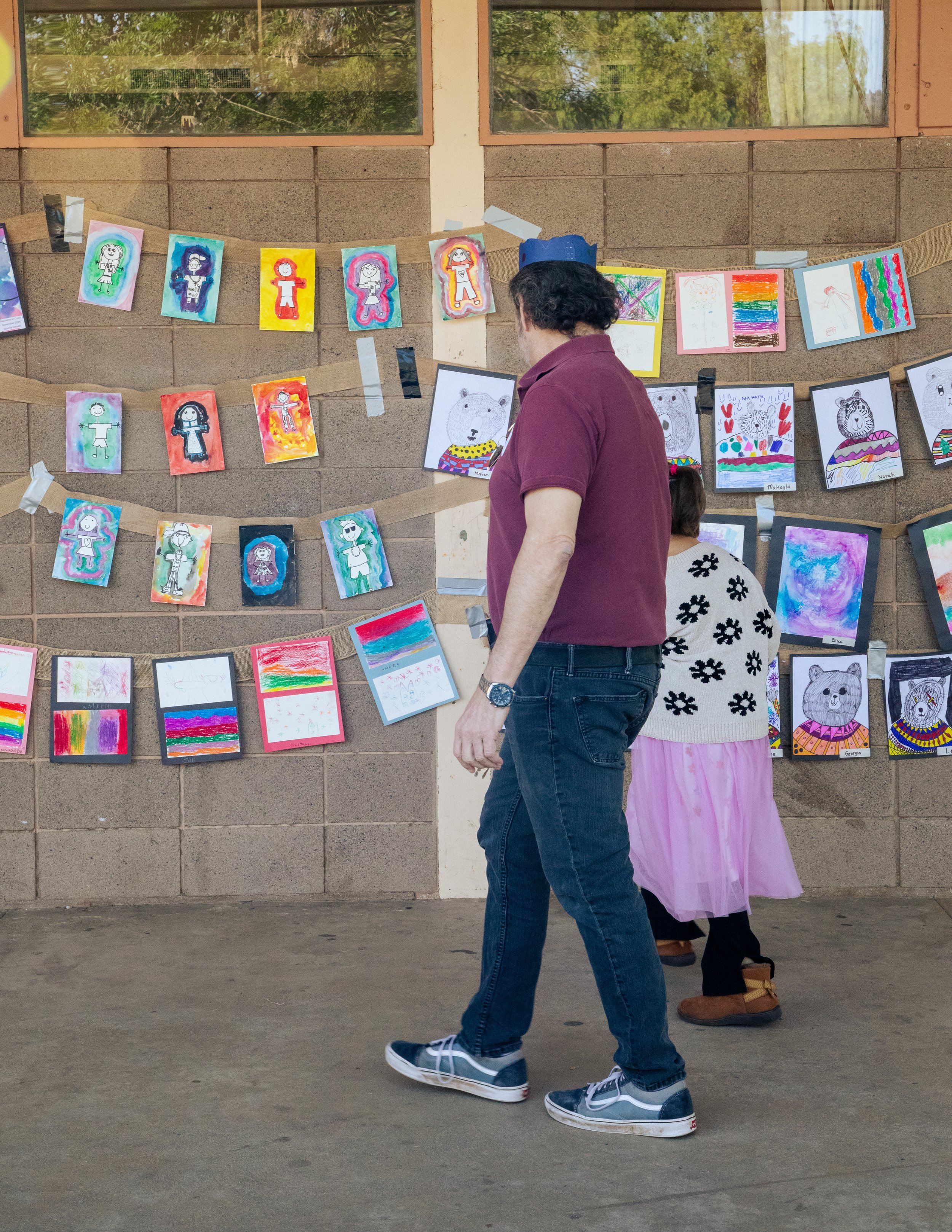 Children and an adult walking past a display of colorful children's artwork hanging on a wall outdoors.
