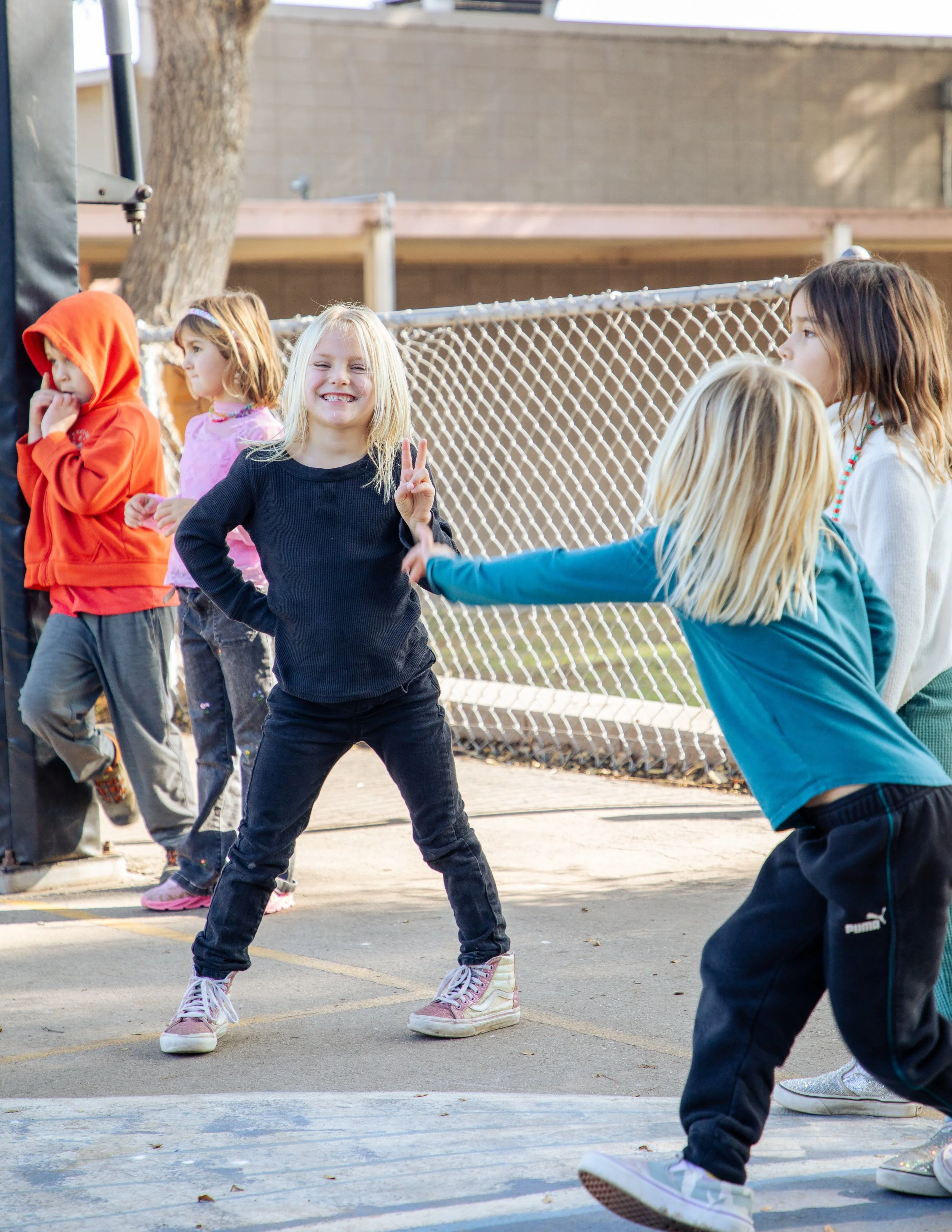 A group of children standing in a line outdoors, with one girl in the foreground making a peace sign and smiling, while others wait their turn, near a chain-link fence and a tree.