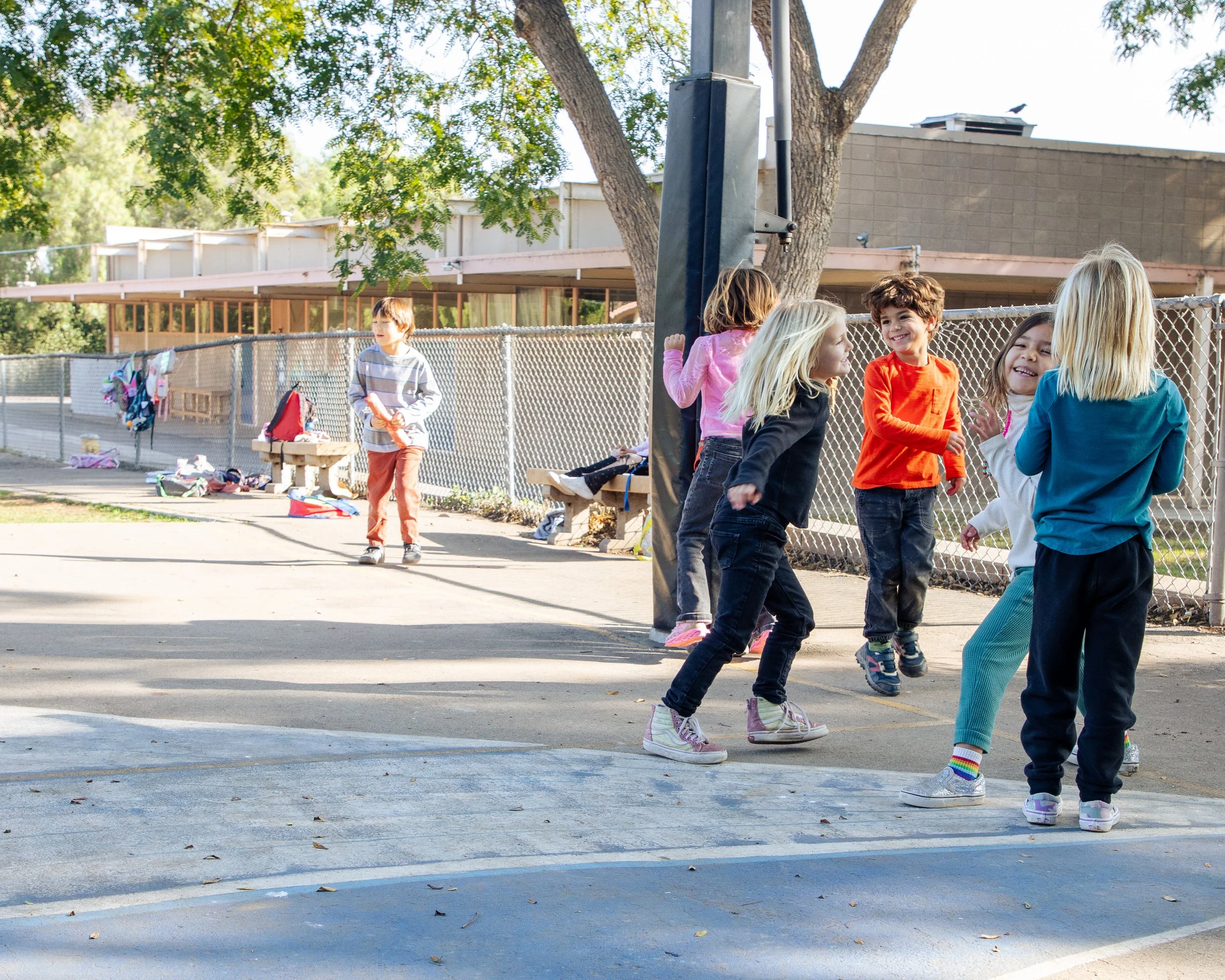 A group of children are playing outside near a chain-link fence on a sunny day. Some children are talking and laughing while others are standing or sitting nearby, with belongings scattered on the ground.