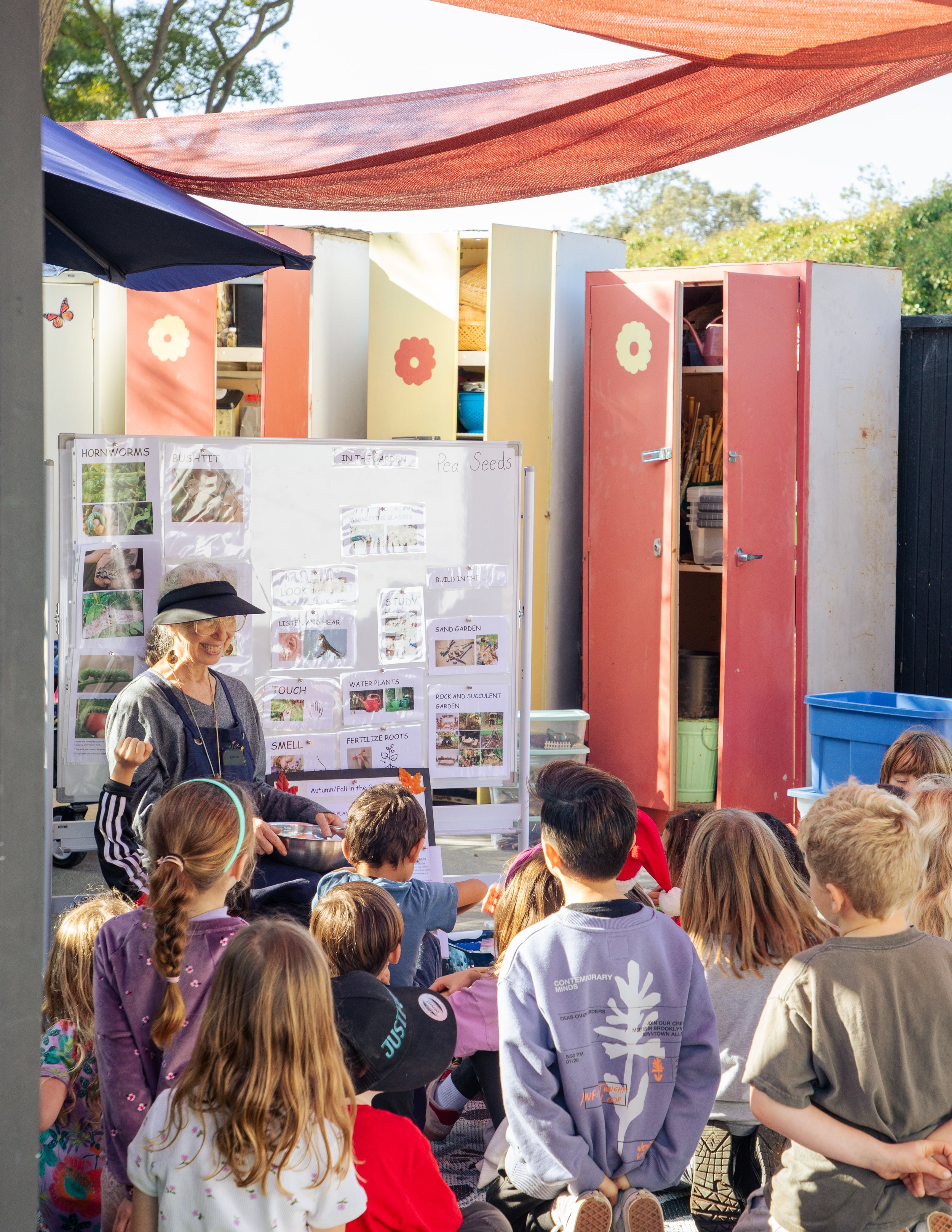 A group of children gathered outdoors, listening to a woman who is demonstrating or talking about plants or gardening. The woman is smiling, holding a bowl, and stands in front of a white display with pictures and labels related to gardening. Bright sunlight and colorful lockers and shade sails are visible.