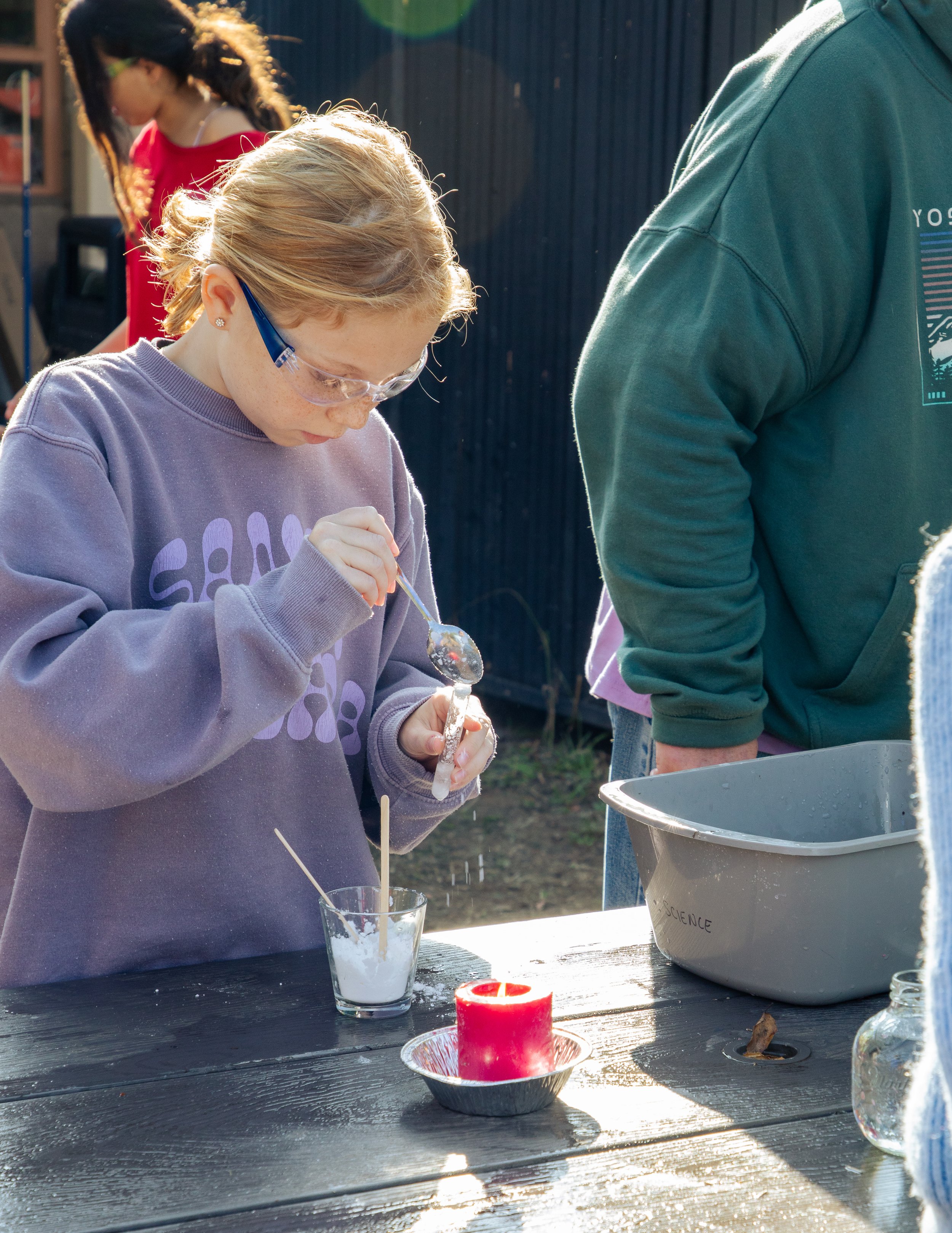 A girl with glasses and a purple sweatshirt is pouring a white liquid into a glass from a small spoon at an outdoor science activity. Other supplies include a candle, a large container labeled 'Science,' a glass jar, and a candle holder on a black wooden table. A person in a green hoodie is nearby, and another girl is in the background.