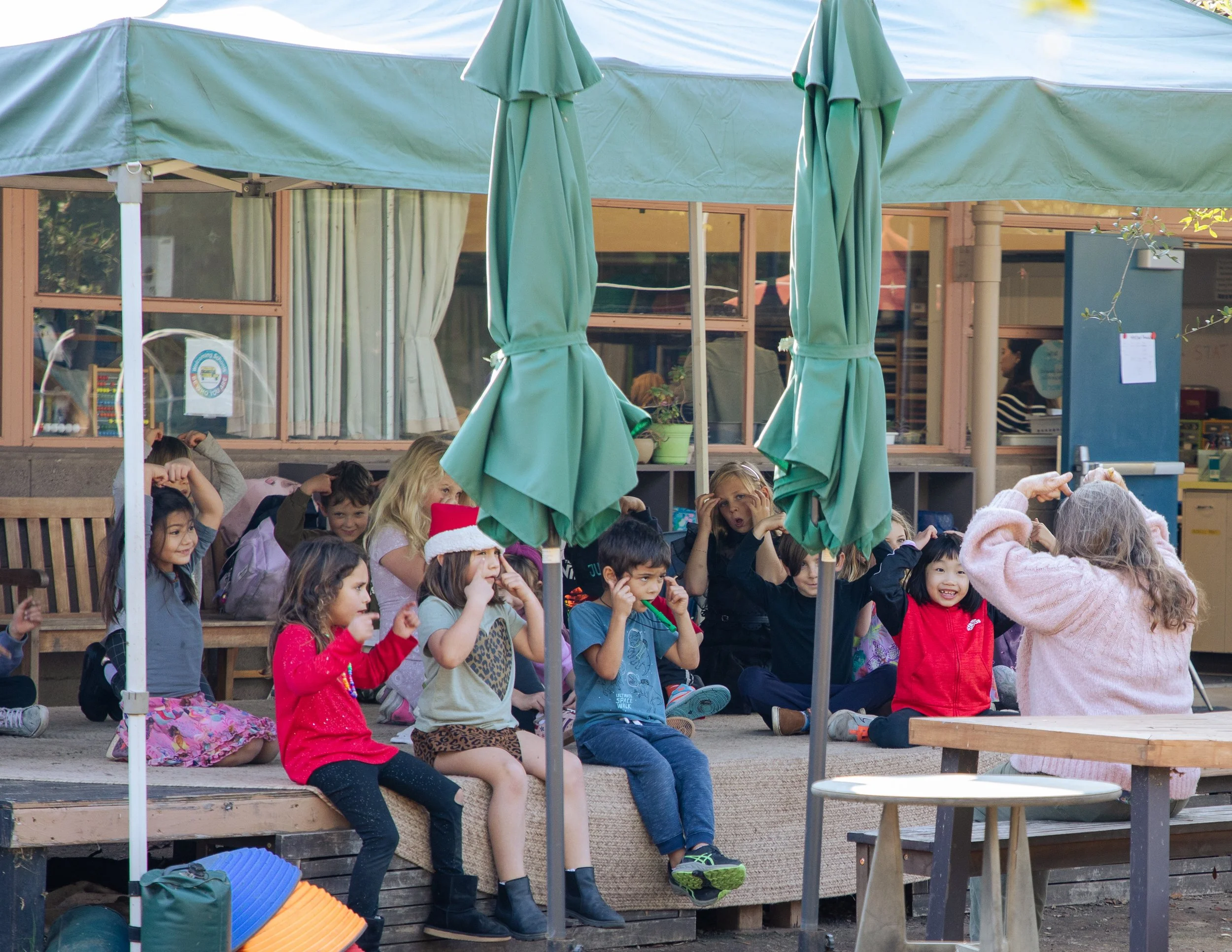 Children sitting on outdoor steps under green umbrellas at school or daycare during daytime.