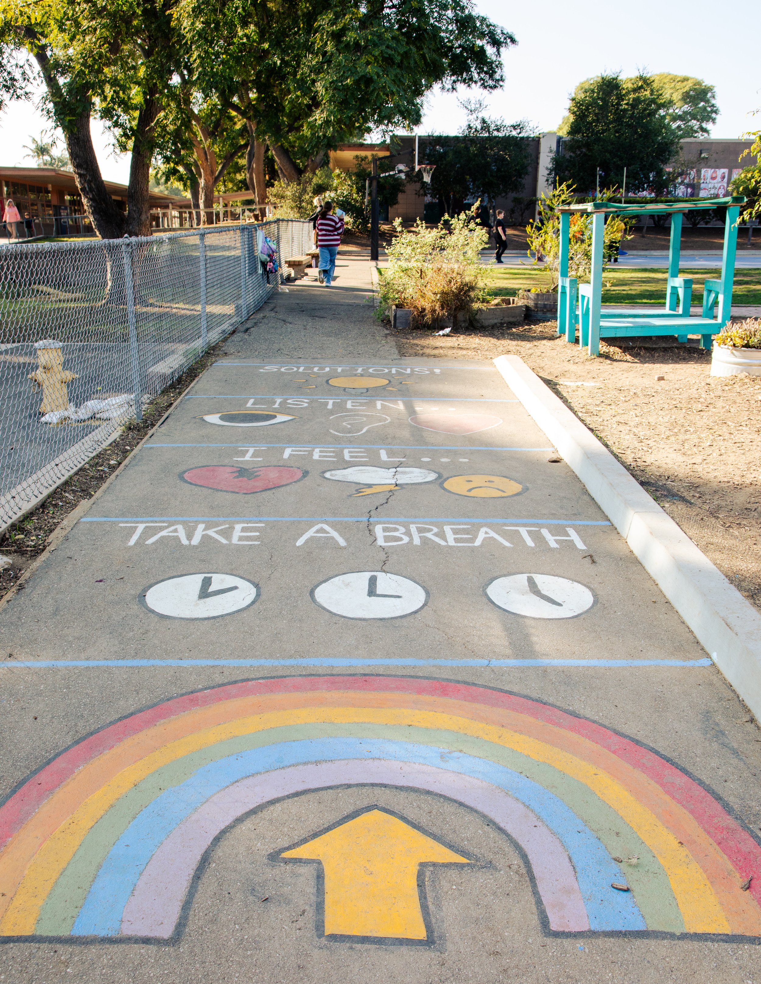Colorful rainbow painted on pavement with motivational phrases and symbols, including a heart and a rainbow, in a park setting.