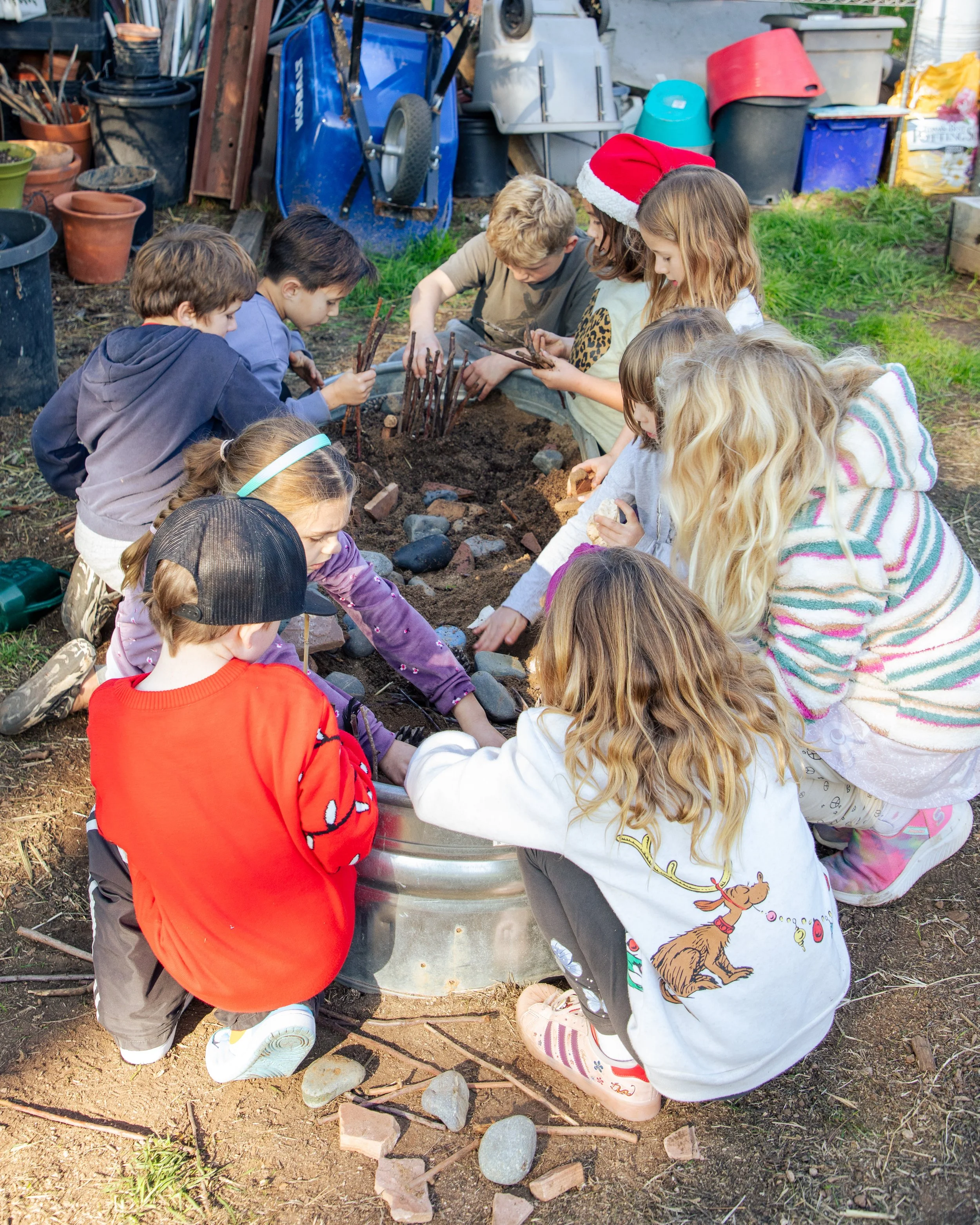 Group of children gathering around a fire pit outdoors, some wearing festive holiday clothing including a Santa hat, and arranging rocks and sticks around the fire pit for a campfire or outdoor activity.