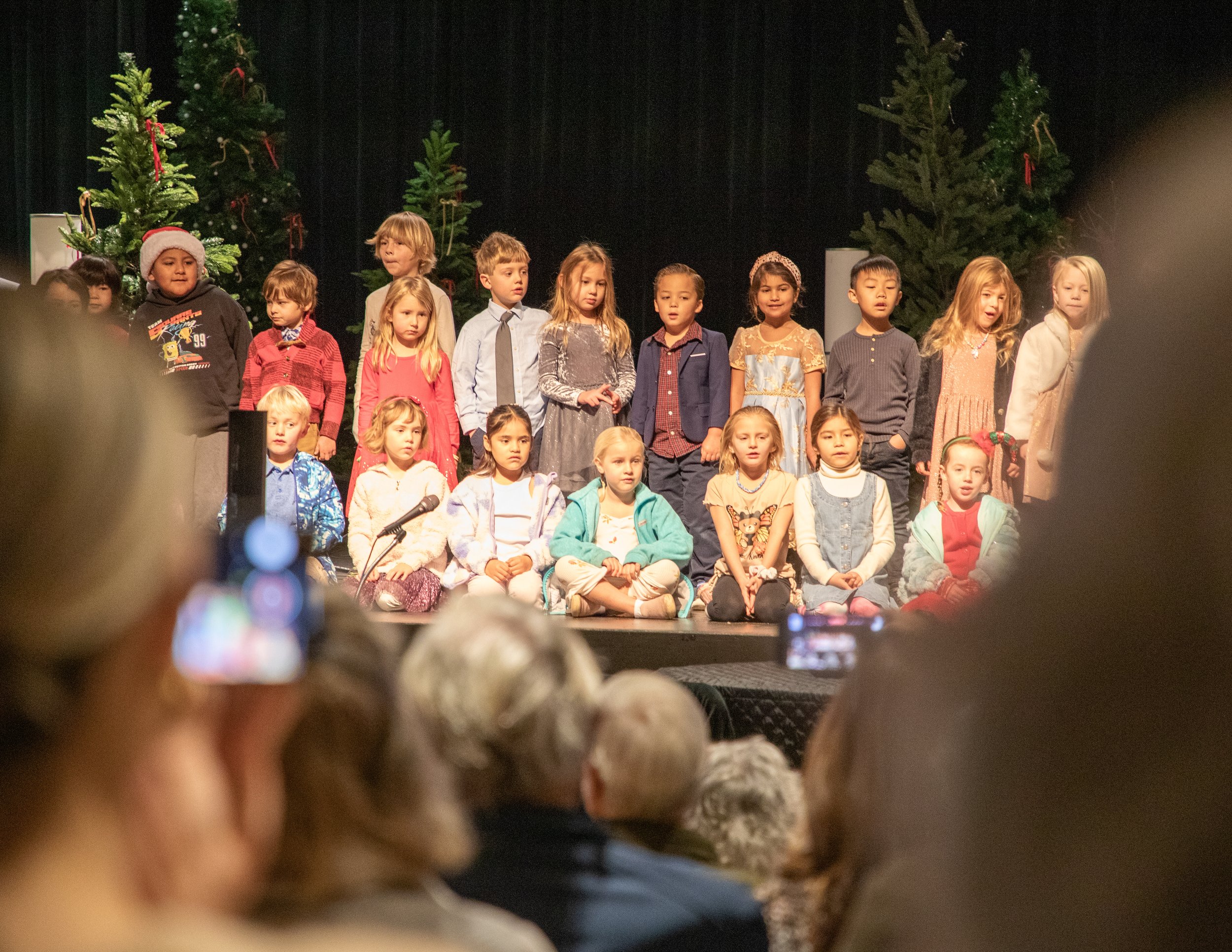 Children standing on stage during a Christmas or holiday performance with decorated Christmas trees in the background.
