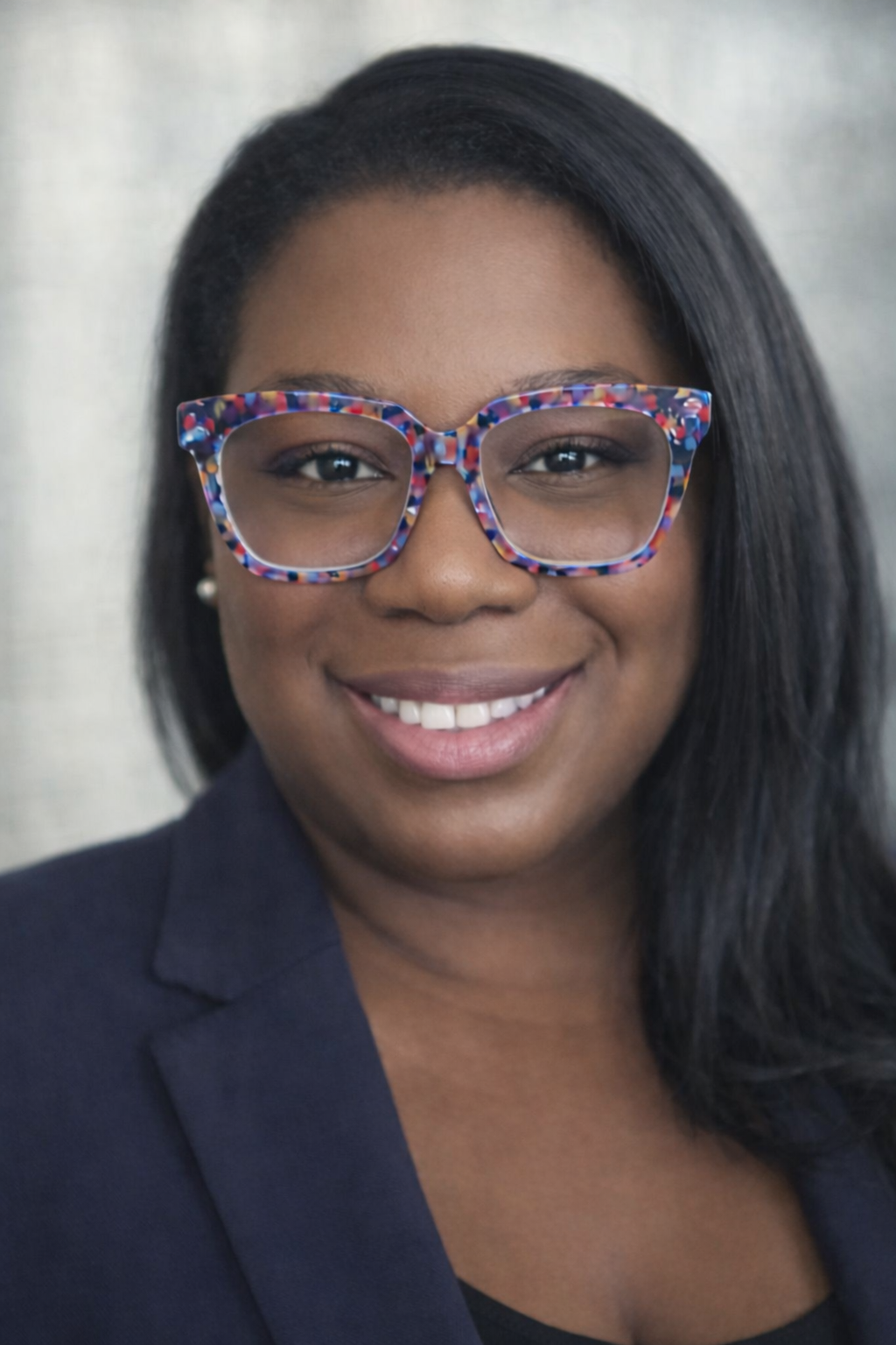 A smiling woman wearing colorful, patterned glasses and pearl earrings, dressed in a navy blazer.