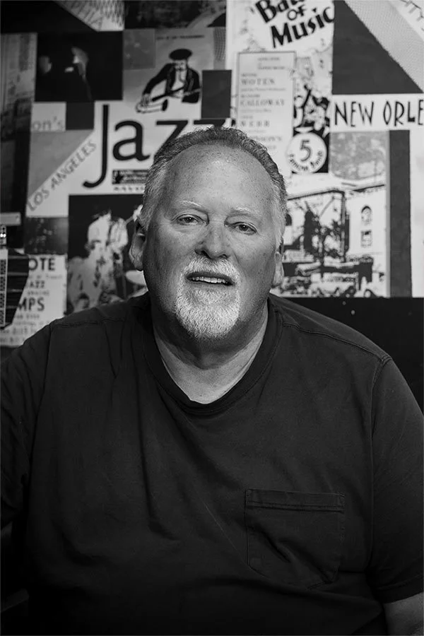 A middle-aged man with a beard and short hair sitting in front of a collage of music and city posters in black and white.