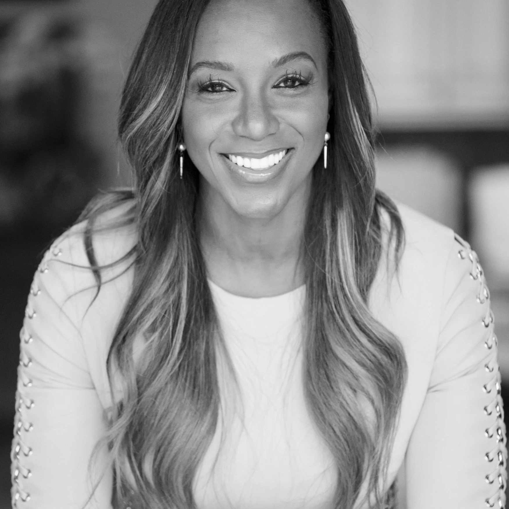 A smiling woman with long, wavy hair, wearing earrings and a light-colored top with embellishments on the shoulders.
