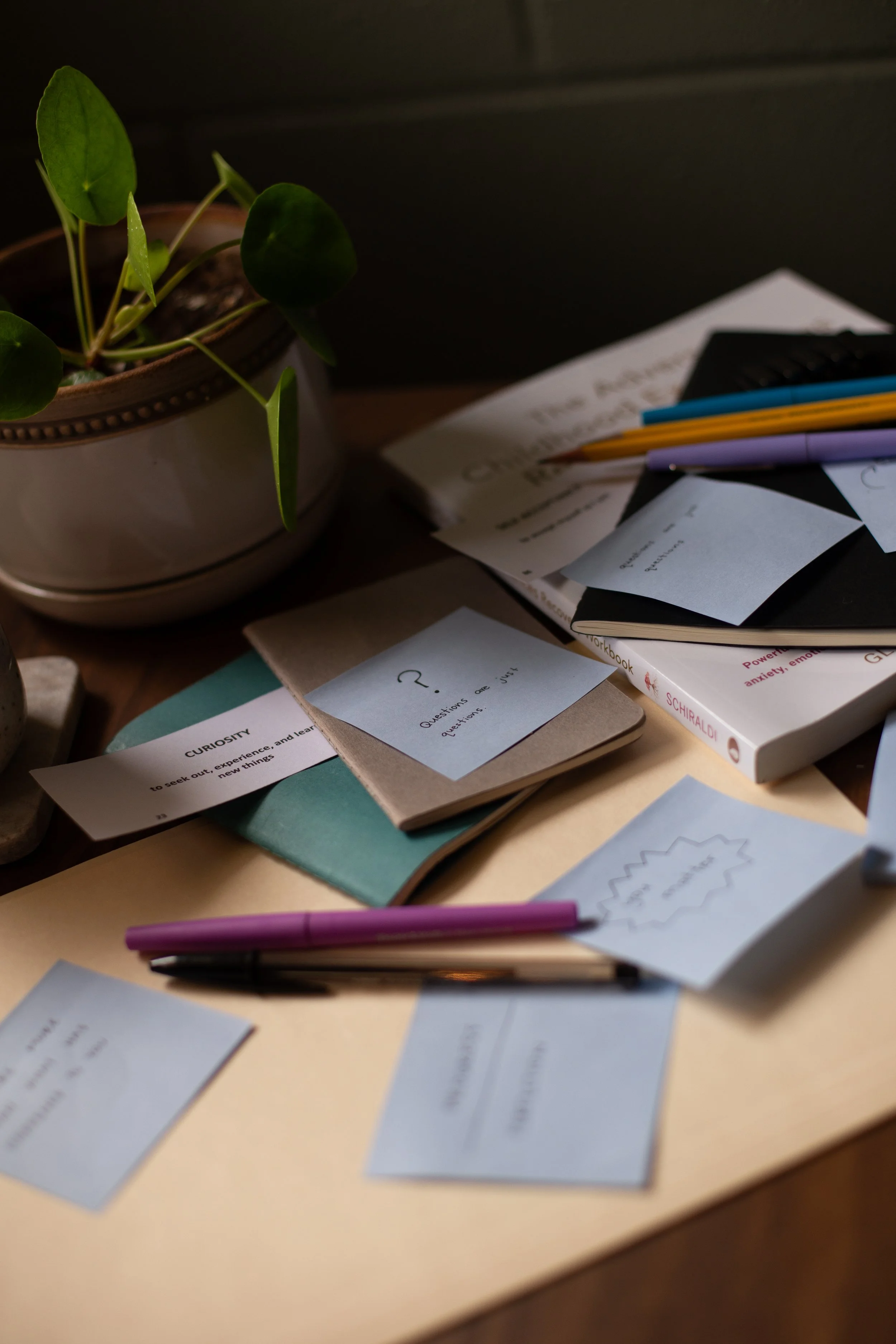 A desk with scattered notes and papers, including a handwritten question mark on a post-it note, a purple pen, a black notebook, and a potted plant with green leaves.