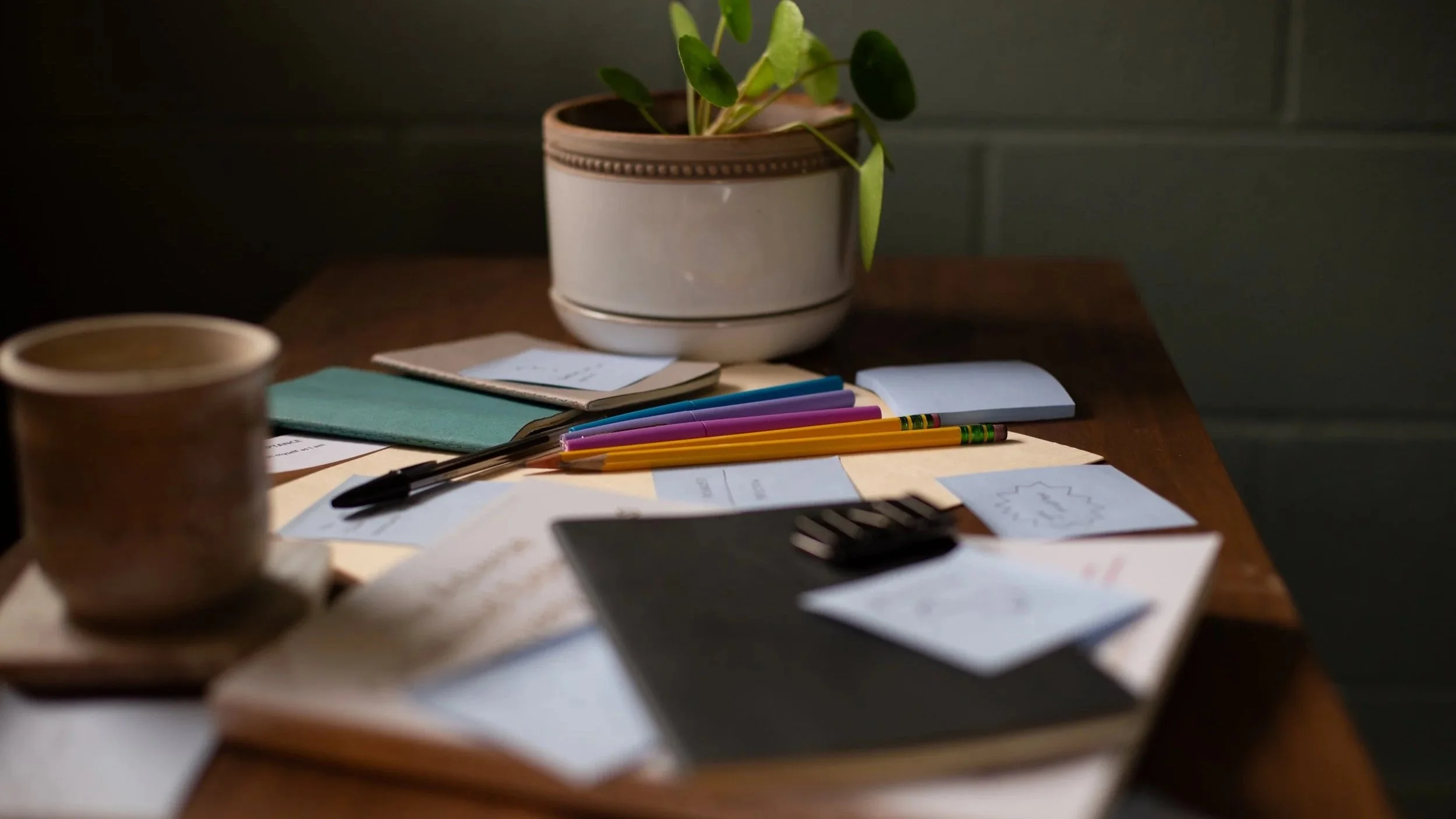 A wooden desk with colored pens, notebooks, papers, and a potted plant, illuminated softly, with a coffee cup partially visible in the foreground.