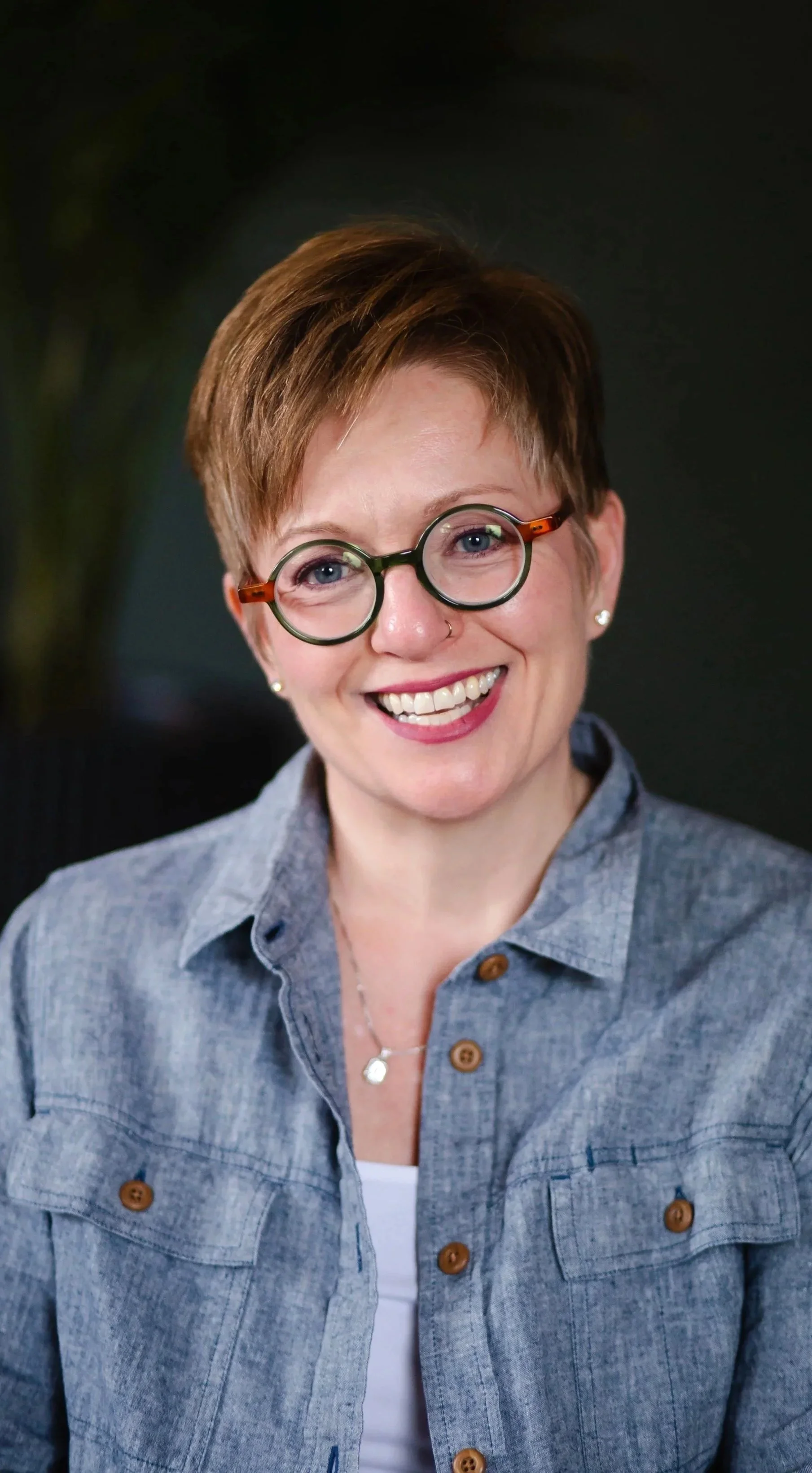 A woman with short red hair and glasses smiling, wearing a denim shirt and a necklace, with a dark background.