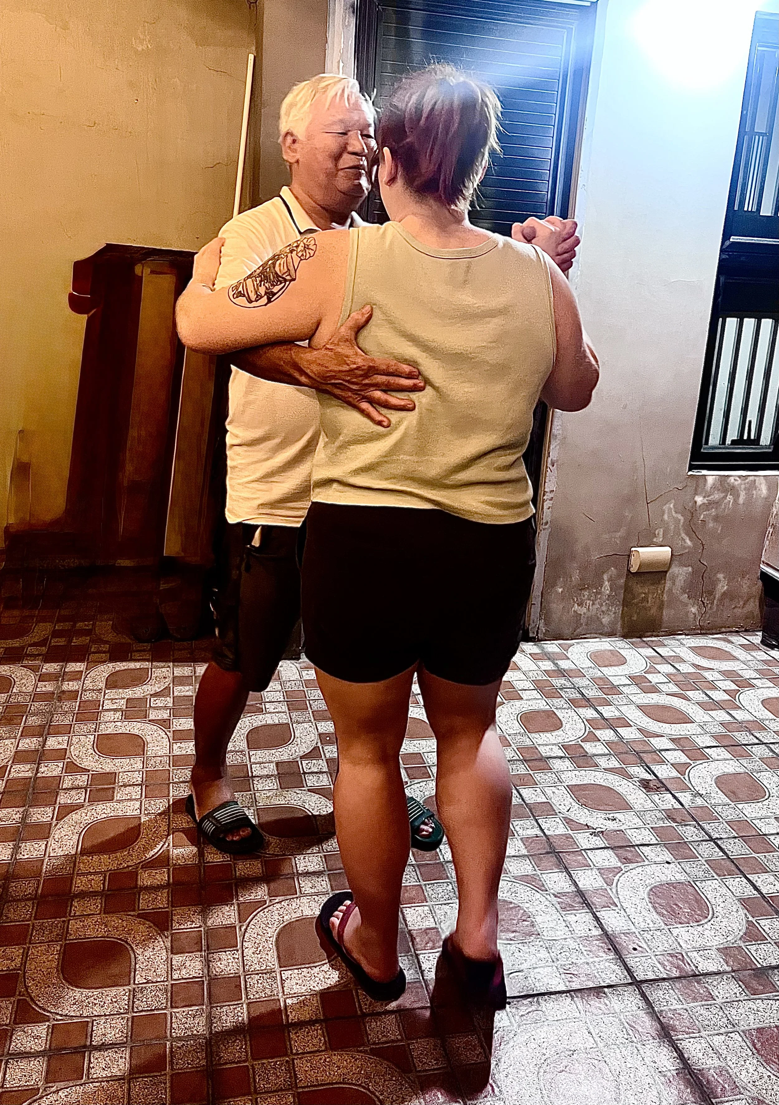 A man and a woman dancing closely in a room with tiled floor and wooden walls, with open windows letting in light.