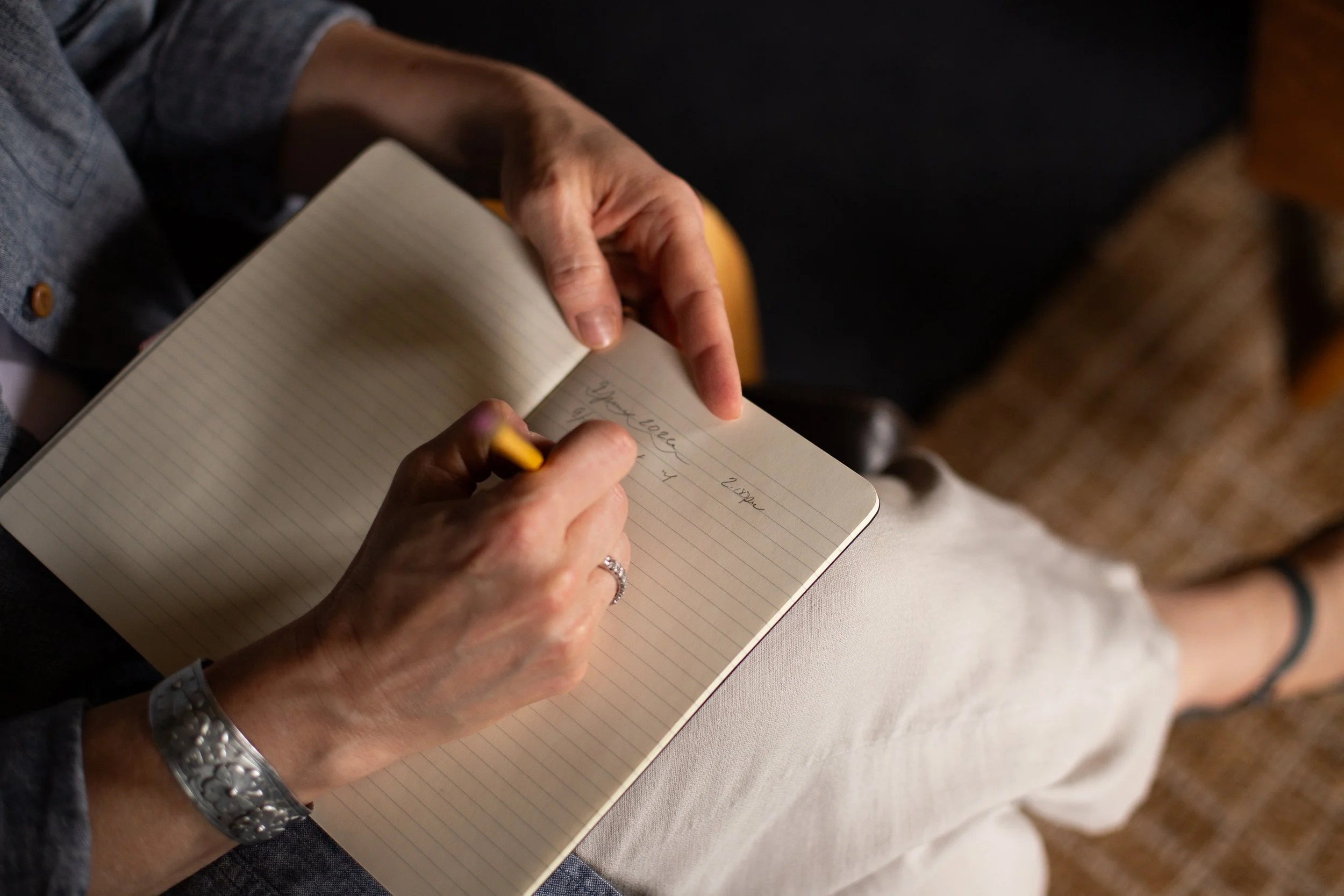 Close-up of a person writing in a lined notebook with a yellow pen, with another person holding the notebook. The setting appears to be indoors.