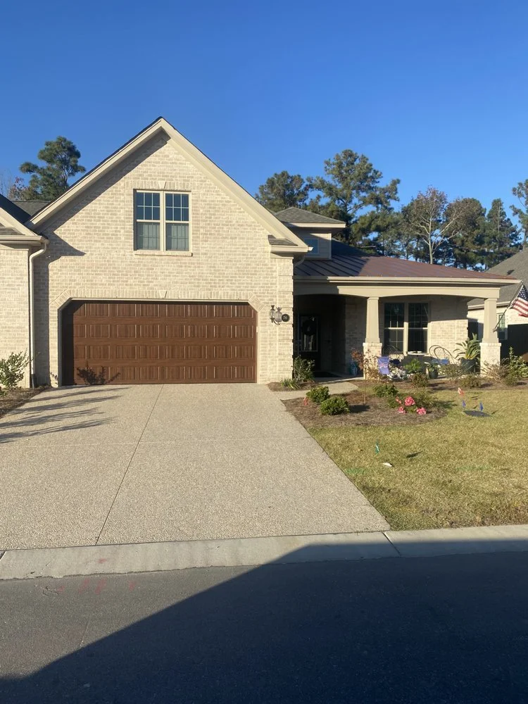 A modern suburban house with a brick exterior, a two-car garage with a brown door, a small front porch with columns, and a well-maintained lawn with some small plants and pink flowers.
