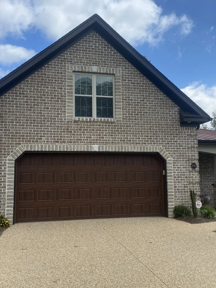 Front view of a brick house with a closed dark brown garage door, window above the garage, and a small garden with plants on the right side.