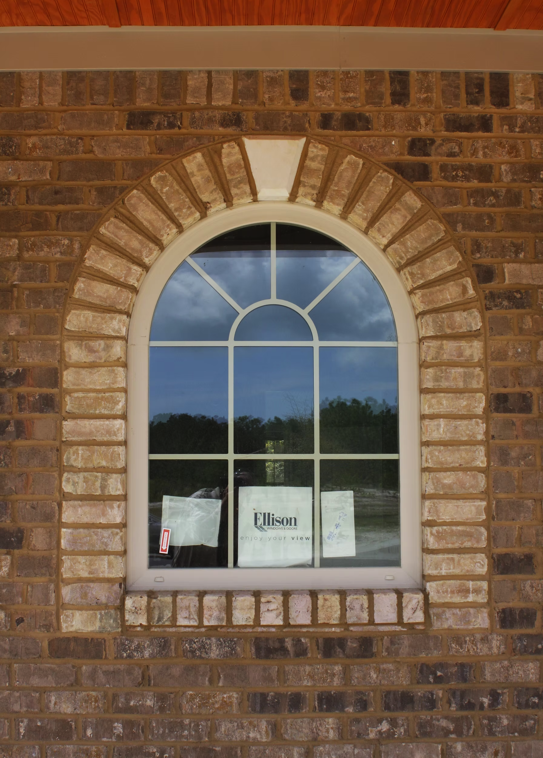 A brick wall with an arched window showing trees and a cloudy sky outside. A sign in the window reads 'Ellison Windows & Doors' with the slogan 'enjoy your view.'