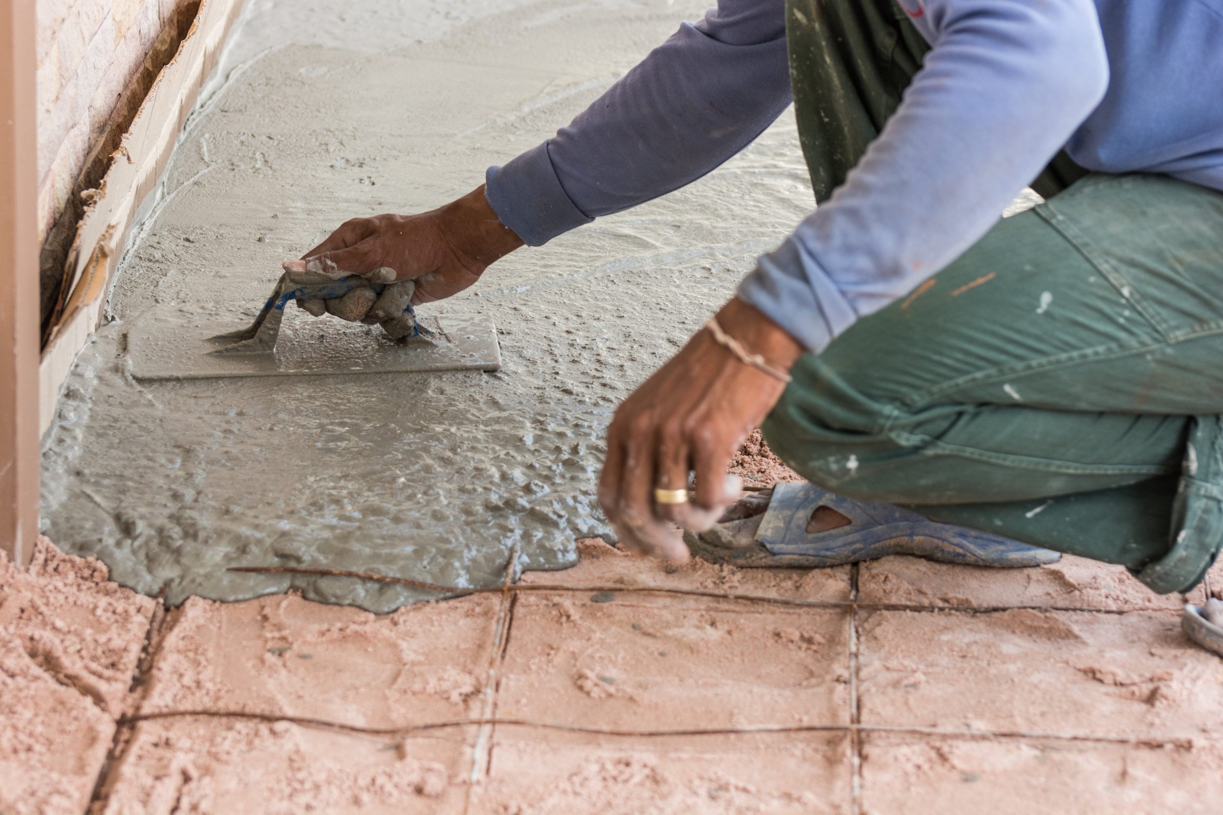 Person kneeling down while applying a layer of wet cement to a floor, with exposed brick wall to the side.