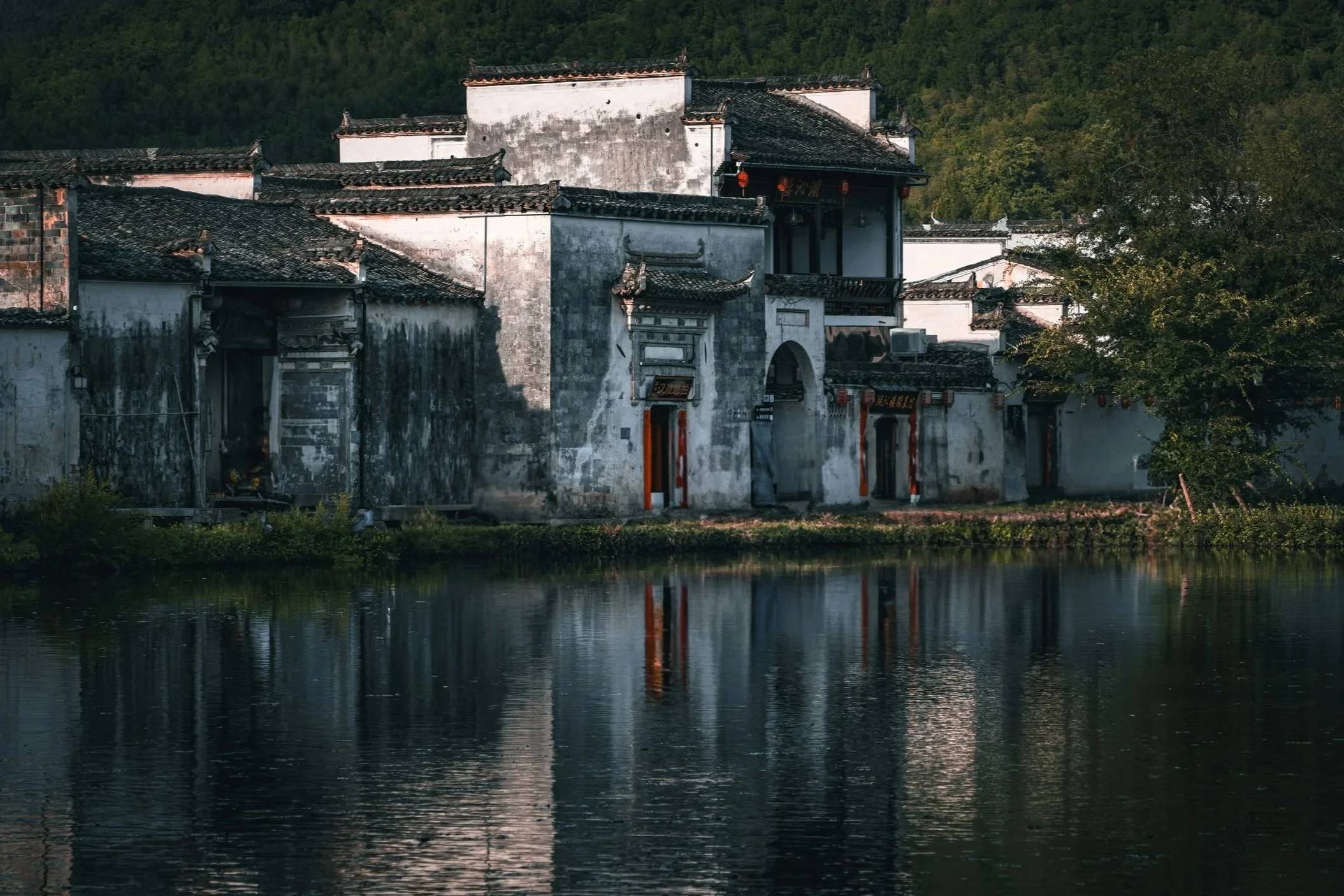 Shexian, Huizhou // Traditional Chinese white-walled buildings with black-tiled roofs beside a body of water, surrounded by trees and green hills.