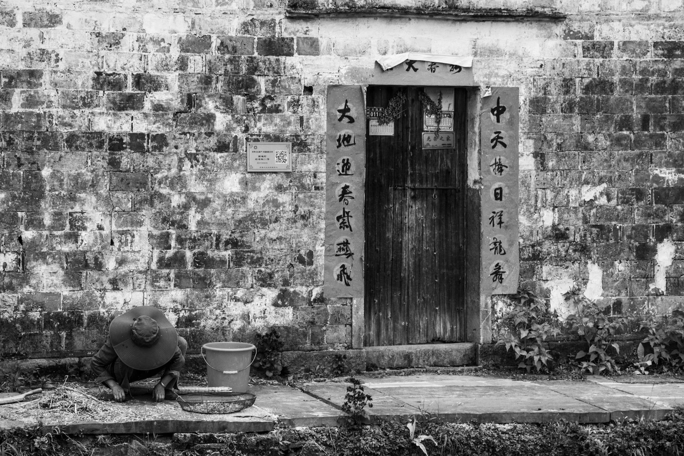 A person wearing a wide-brimmed hat watering plants in front of a brick wall with a wooden door and Chinese calligraphy on paper scrolls.