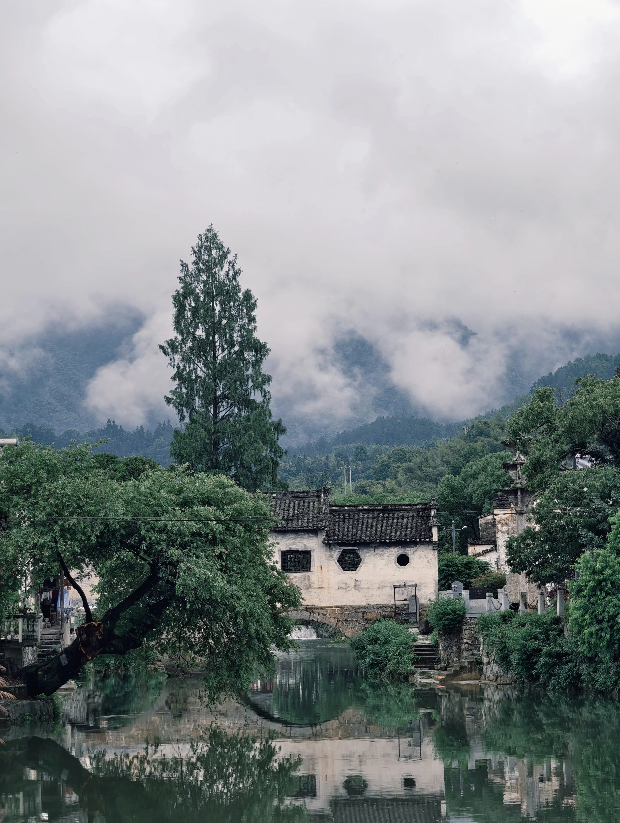A tranquil river flows through a traditional village with white buildings and dark tiled roofs, surrounded by lush green trees and fog-covered mountains in the background, under a cloudy sky.