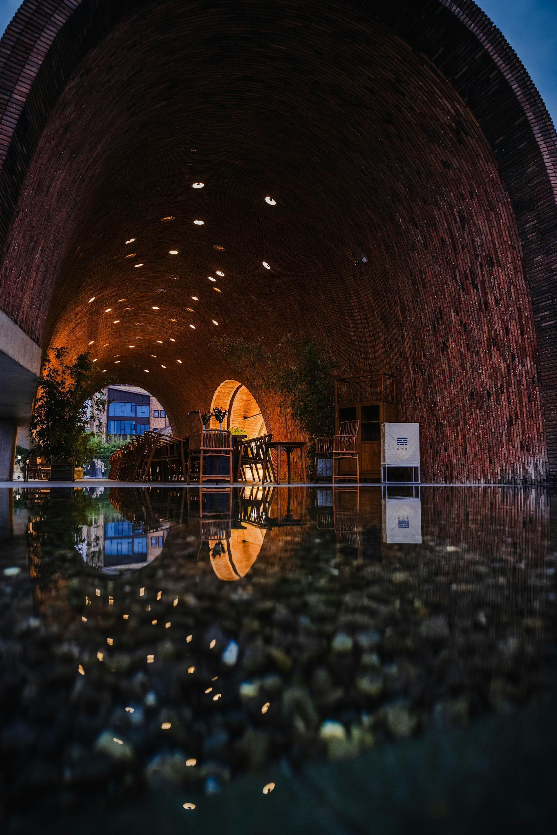 Interior view of a curved brick archway ceiling with small round lights and a reflection of the scene on a wet floor, with outdoor buildings and sky visible through an opening.
