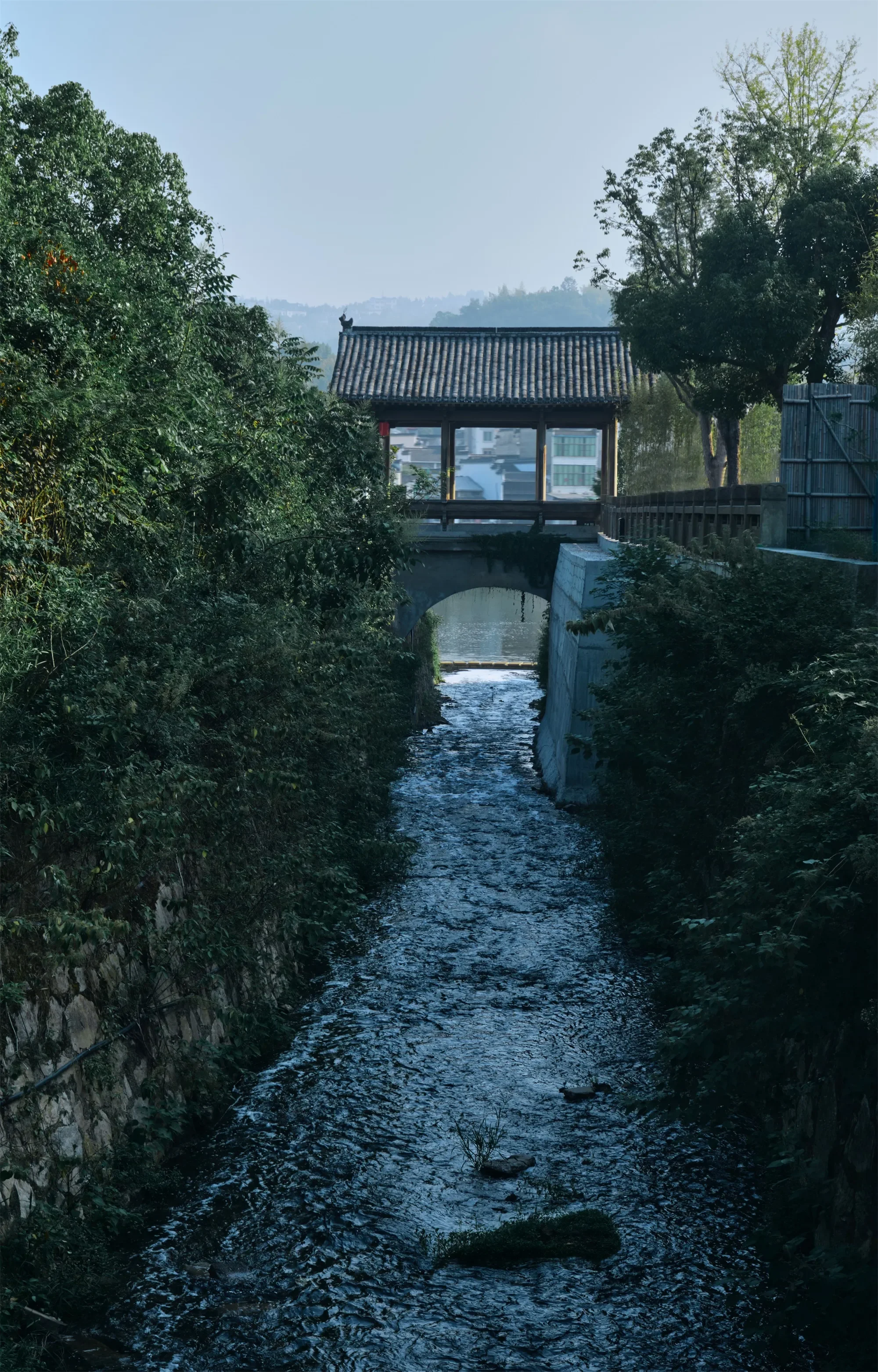 A narrow stream of water flows under a traditional Japanese wooden bridge with tiled roof, surrounded by lush green trees.