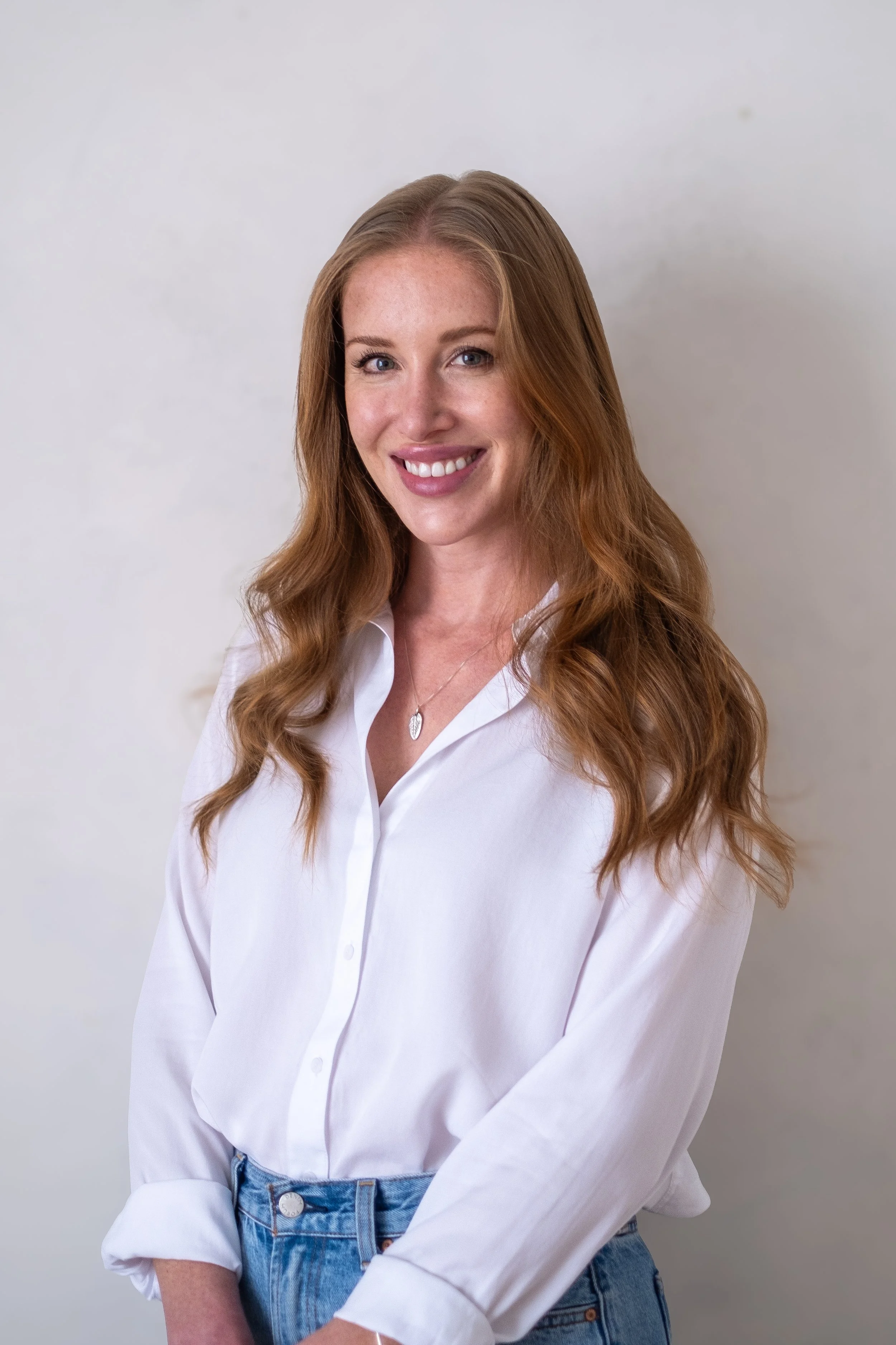 A woman with long, wavy red hair and blue eyes, smiling, wearing a white button-up shirt with the sleeves rolled up and high-waisted blue jeans, standing against a plain light-colored background.