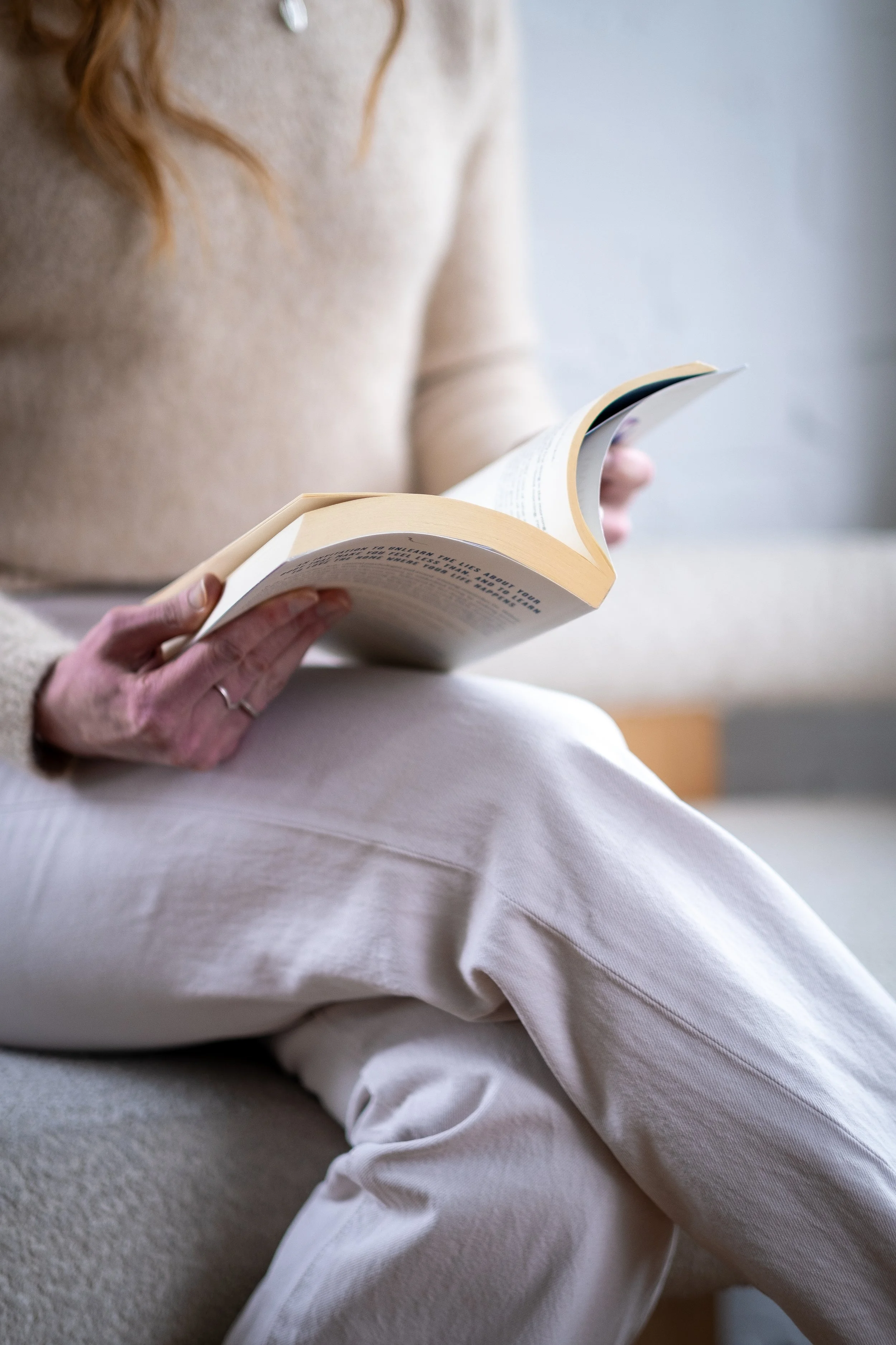 Person sitting on the floor reading a book, wearing a beige sweater and white pants.
