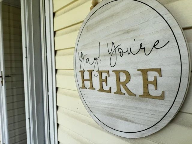 Round wooden sign on a wall that says "Yay! You're HERE" in black cursive and gold block letters.