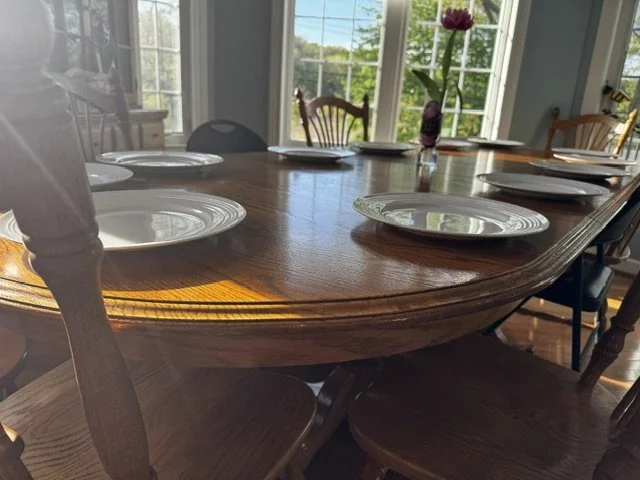 Dining table set with six white plates and a purple flower in a vase, in a sunlit room with large windows and greenery outside.