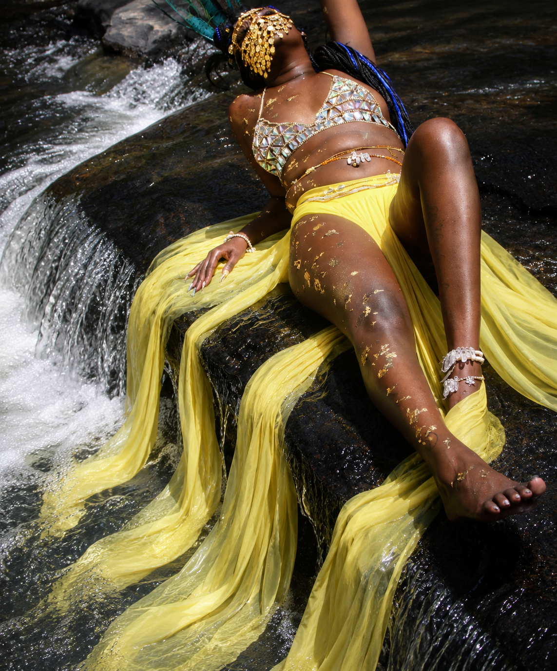 A woman dressed in a rhinestone top, yellow flowing skirt, and gold jewelry, lying on a rock in a stream with water flowing over her body.