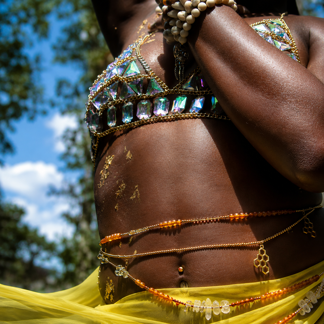 Close-up of a person with dark skin wearing layered, colorful jewelry and golden body paint, outdoors on a sunny day with trees and a blue sky in the background.