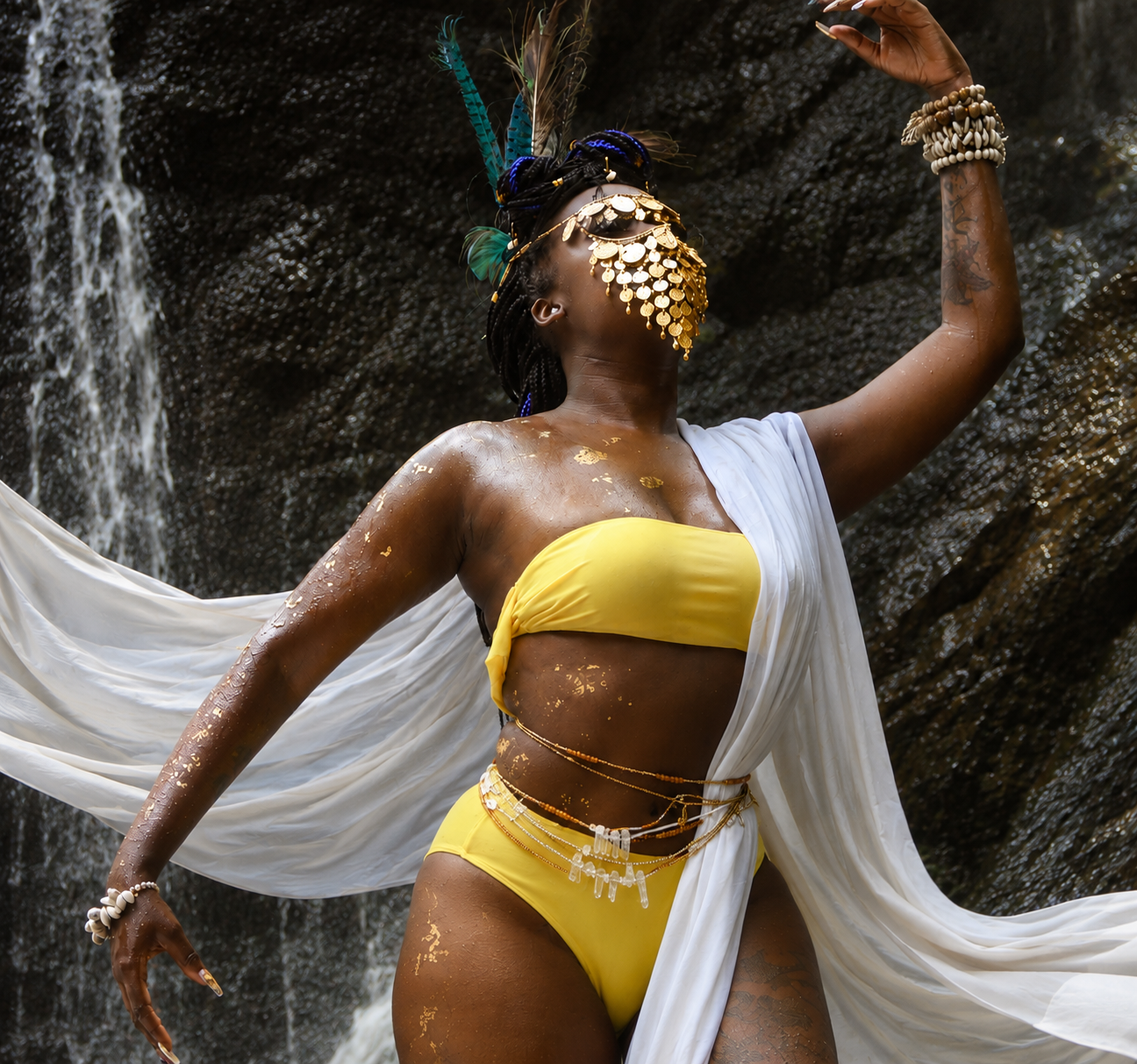 A woman dressed in yellow and white, adorned with gold jewelry, striking a pose in front of a waterfall.