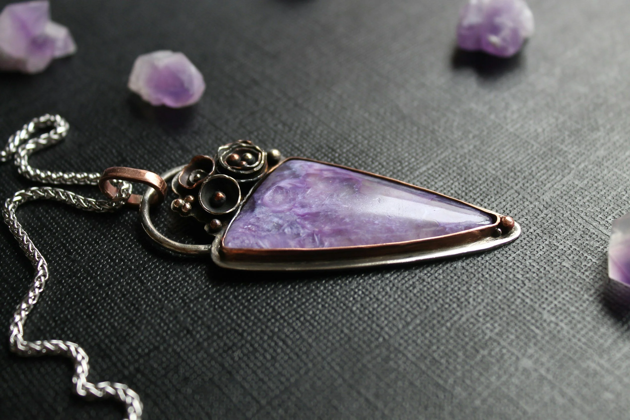 A necklace with a large purple gemstone pendant lying on a black textured surface, with small purple stones scattered around.