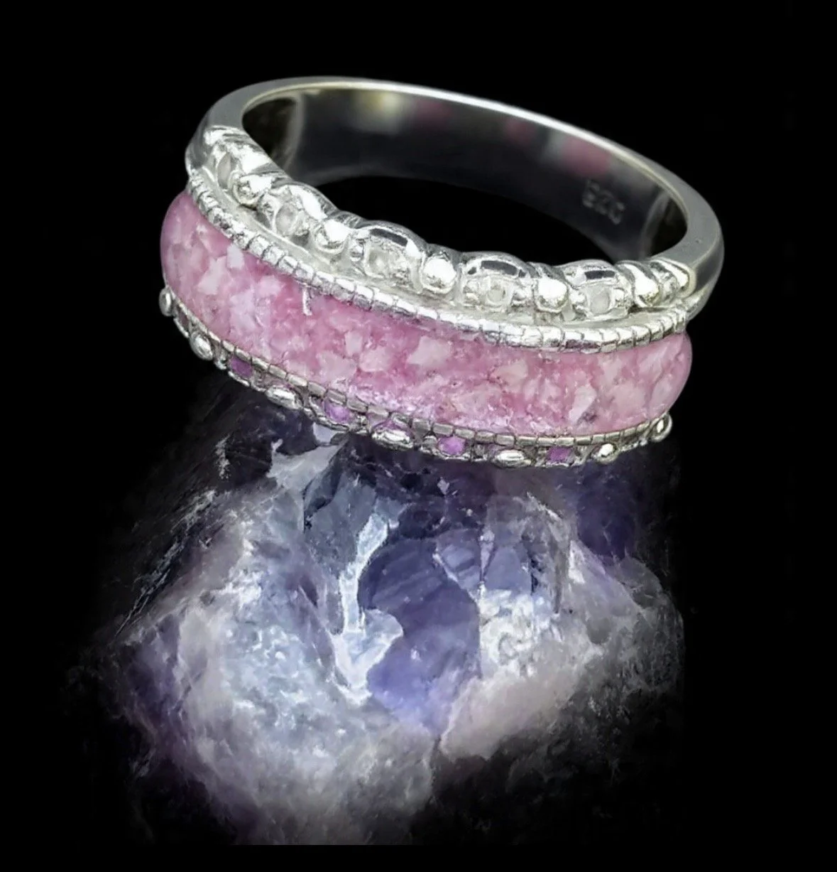 A silver ring with pink resin and ashes and decorative elements, reflecting on a purple gemstone surface.