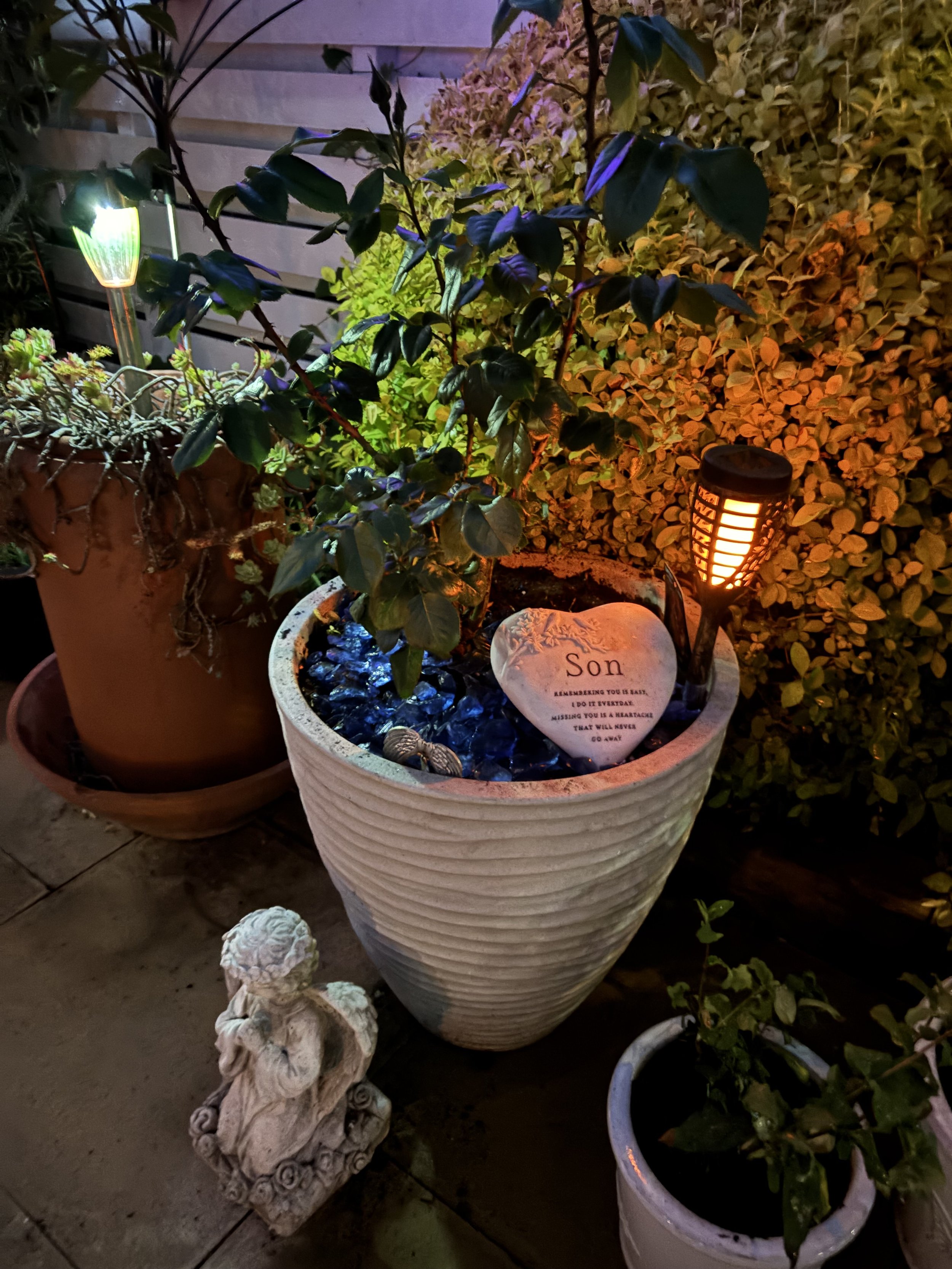 Nighttime garden scene with illuminated pots containing plants and decorative stone sculptures. One pot has a small heart-shaped stone with an inscription, surrounded by blue decorative stones, with a small angel sculpture nearby. The backdrop includes a wooden fence and a hedge.