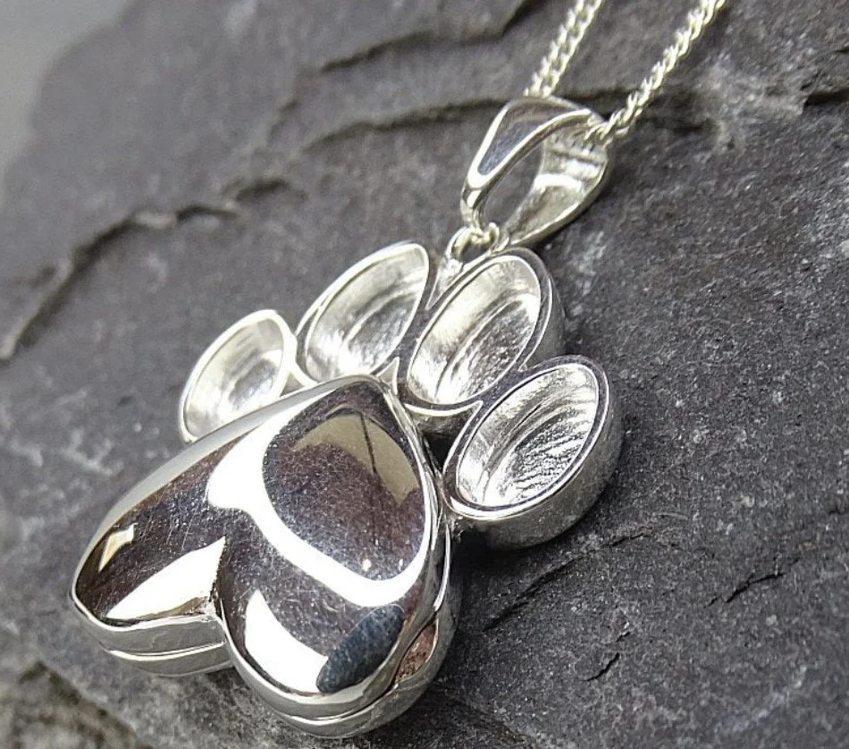 Silver pendant necklace with a paw print design ready for your ashes and resin, resting on a dark surface.