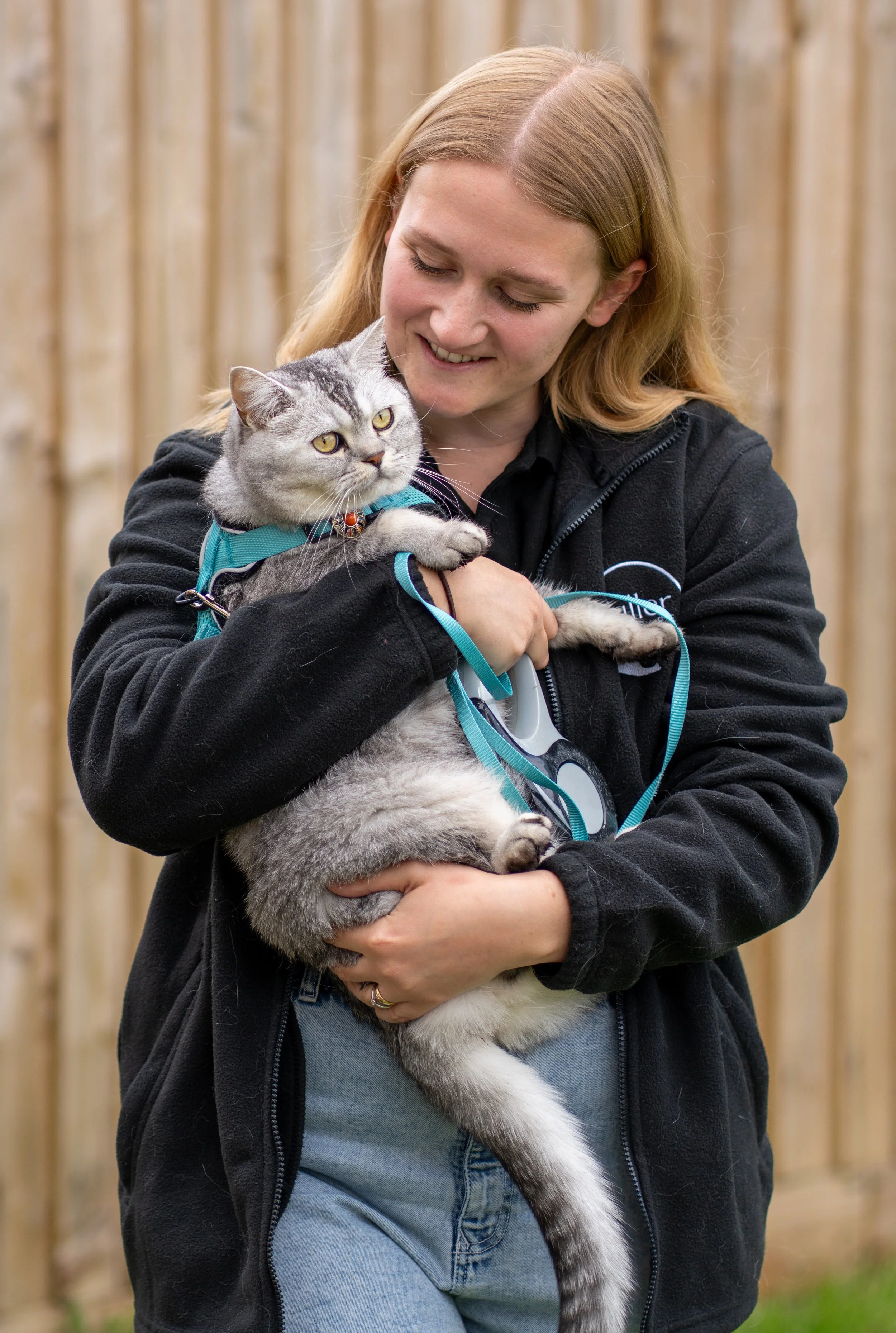 A woman with red hair holding a gray tabby cat with yellow eyes and a blue harness, standing outdoors in front of a wooden fence.