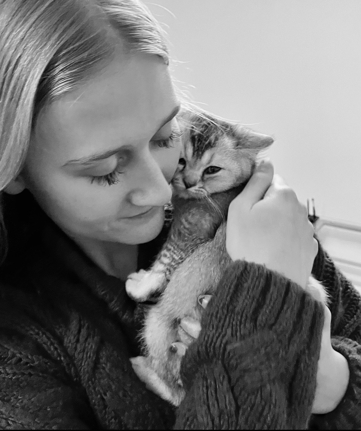 A woman with long hair holding and cuddling a young tabby kitten with closed eyes, both appearing to enjoy a close moment in a cozy indoor setting.