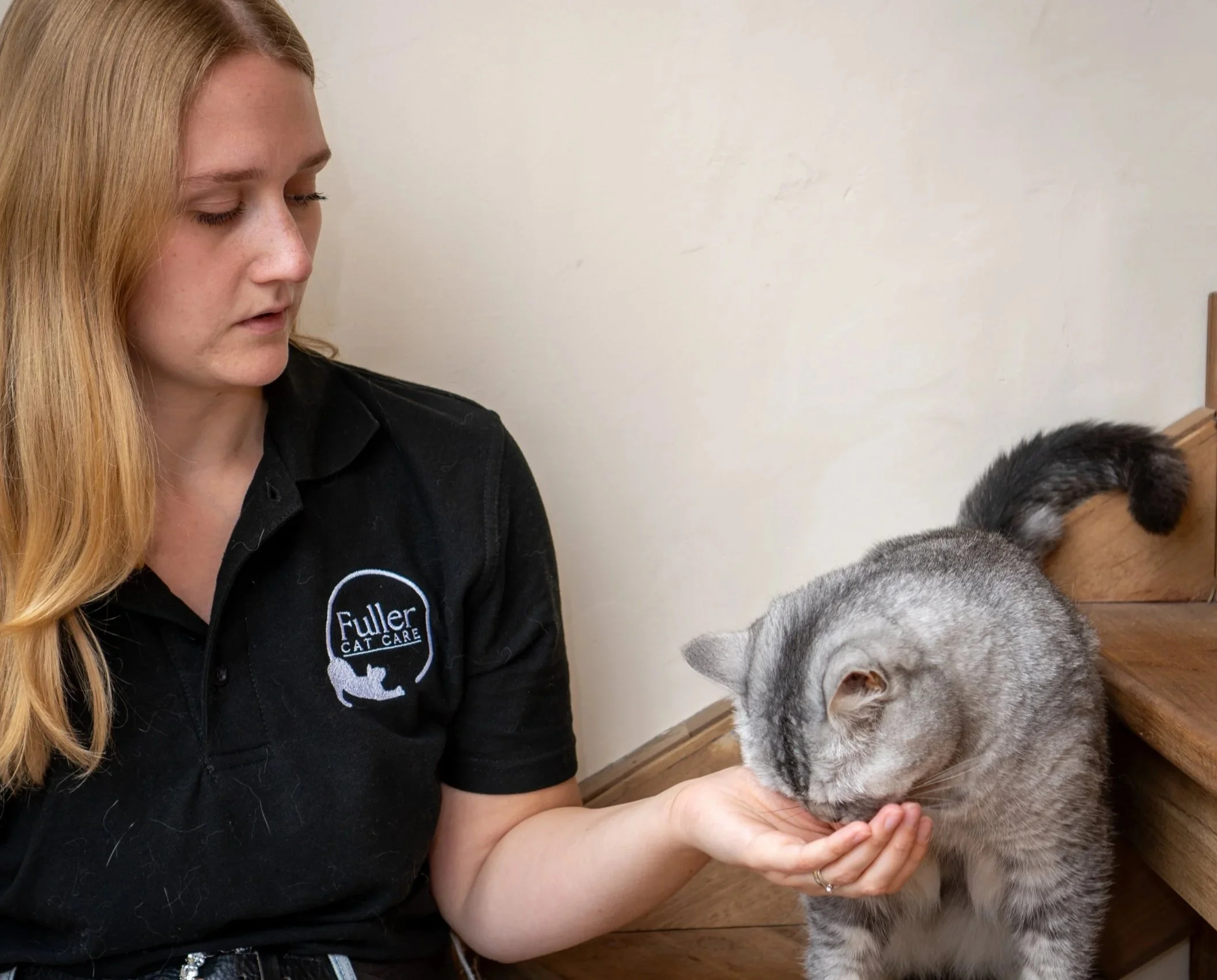 A woman with long blonde hair wearing a black shirt with a Fuller Cat Care logo, gently holding and petting a gray and white Siberian Husky dog. They are indoors against a plain off-white wall.