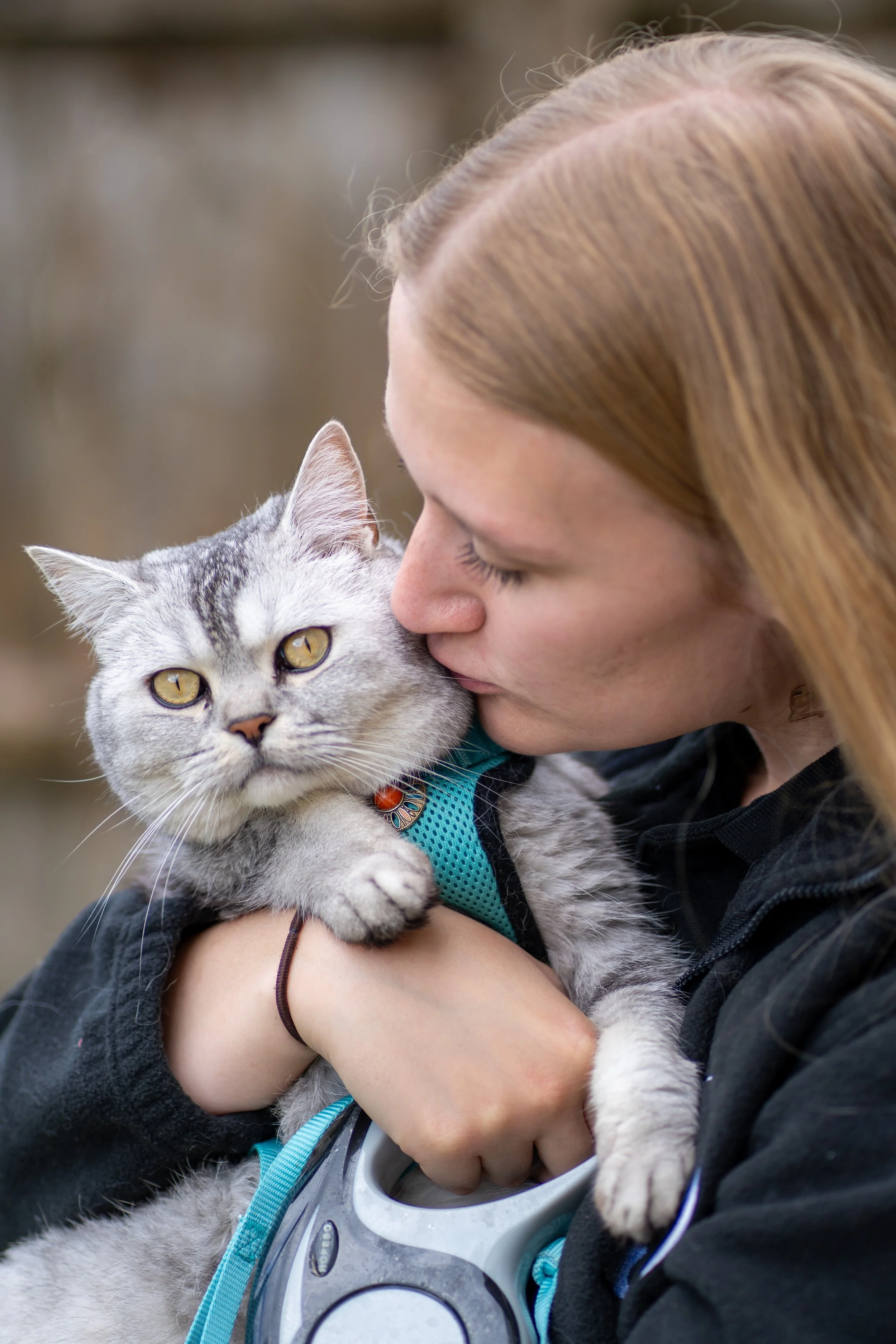 A woman with red hair holding a gray tabby cat close to her face, gently pressing her forehead against the cat's head, outdoors with blurred background.