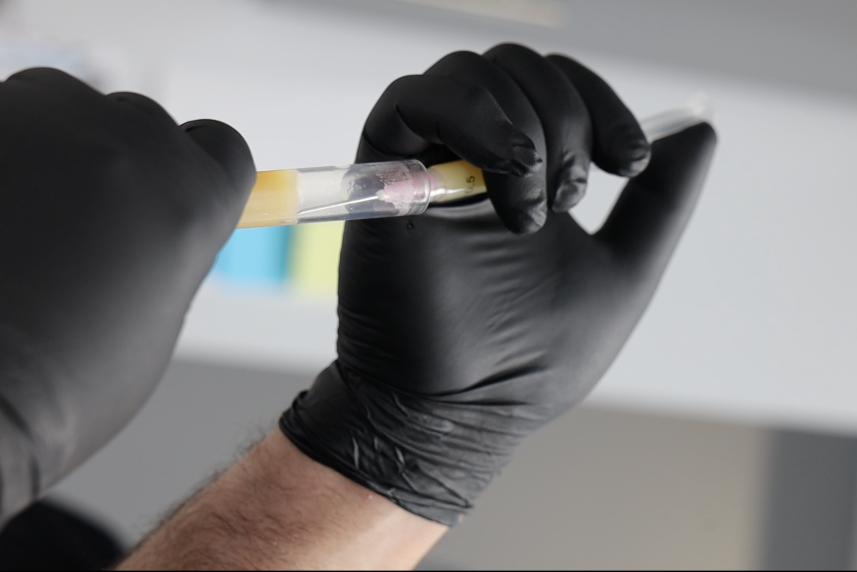 Person wearing black gloves drawing a liquid sample from a test tube.