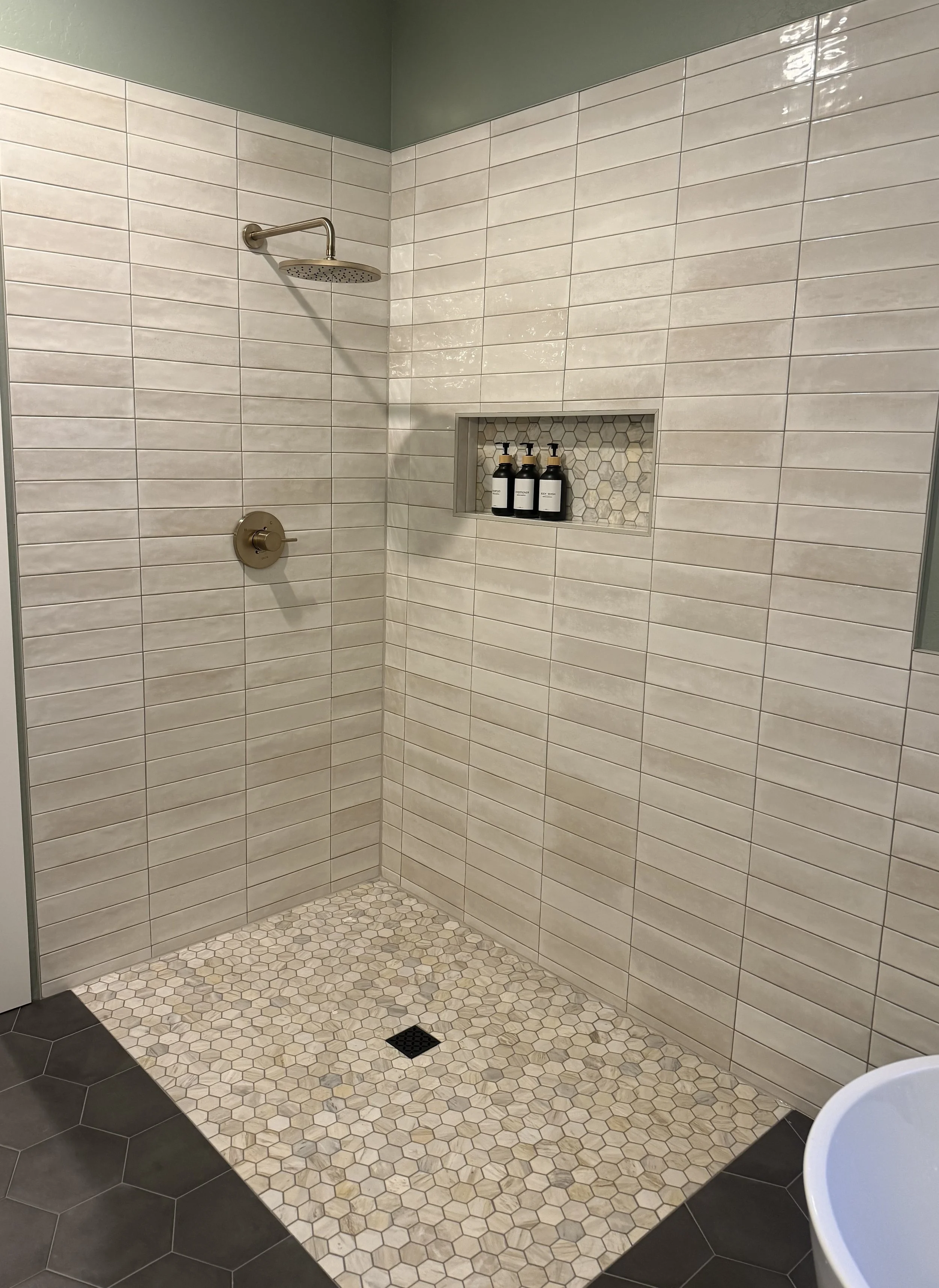 A modern walk-in shower with beige rectangular wall tiles, a hexagonal tiled floor, and a built-in shower niche with three bottles of soap or shampoo. The shower has a rain showerhead and a control knob, with a black drain on the floor.