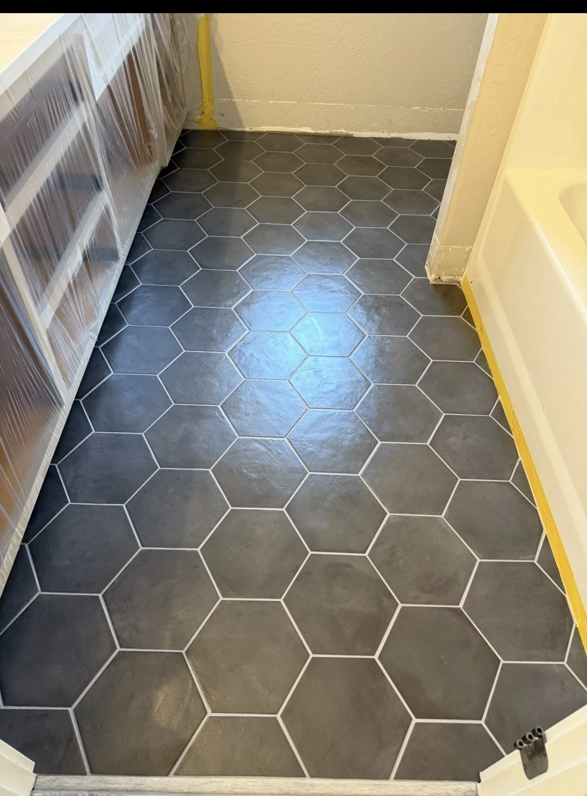 Finished dark gray hexagon tile flooring in a room under construction with beige walls, yellow painter's tape along the baseboards, and some exposed electrical outlets.