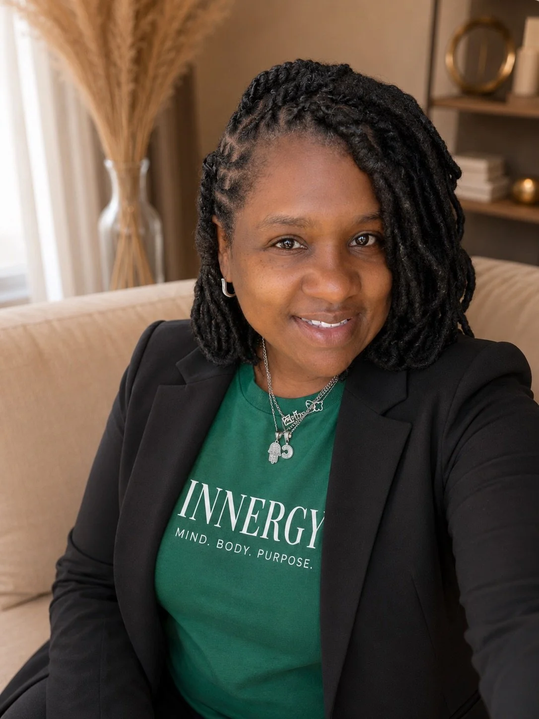 A woman with black styled hair wearing a green 'INNERGY' t-shirt, black blazer, and silver jewelry, sitting indoors on a beige sofa, smiling at the camera.